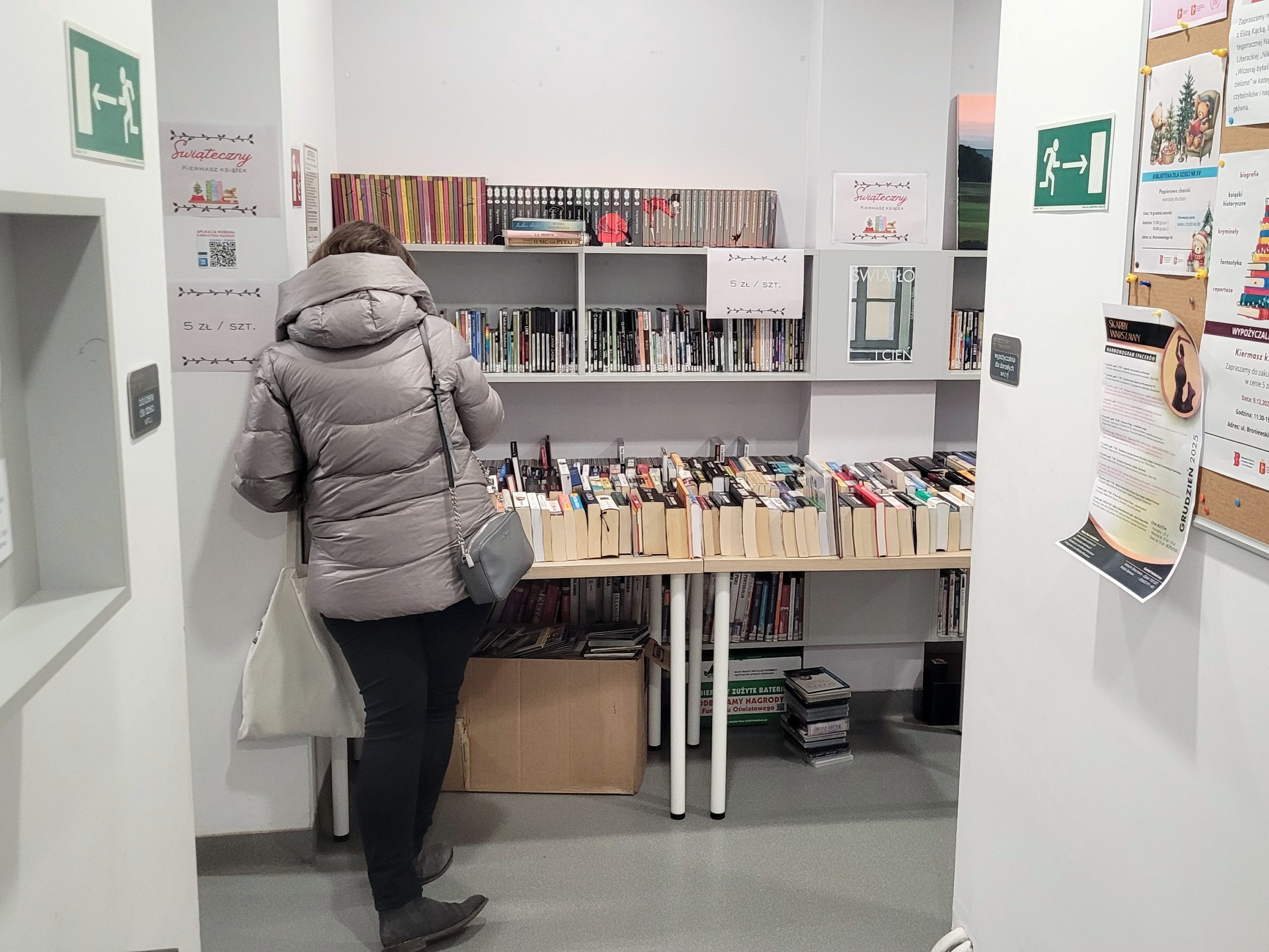 Regały i stoły z używanymi książkami w bibliotece podczas świątecznego kiermaszu; kartki informują o cenie 5 zł za sztukę, z przodu widać osobę przeglądającą książki.
