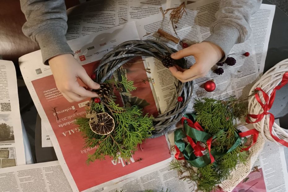 ilustracja graficzna przedstawiająca zdjęcie z relacji zajęć z wieńców zimowych z dziecięcymi dłońmi i świątecznym wieńcem w trakcie jego tworzenia.