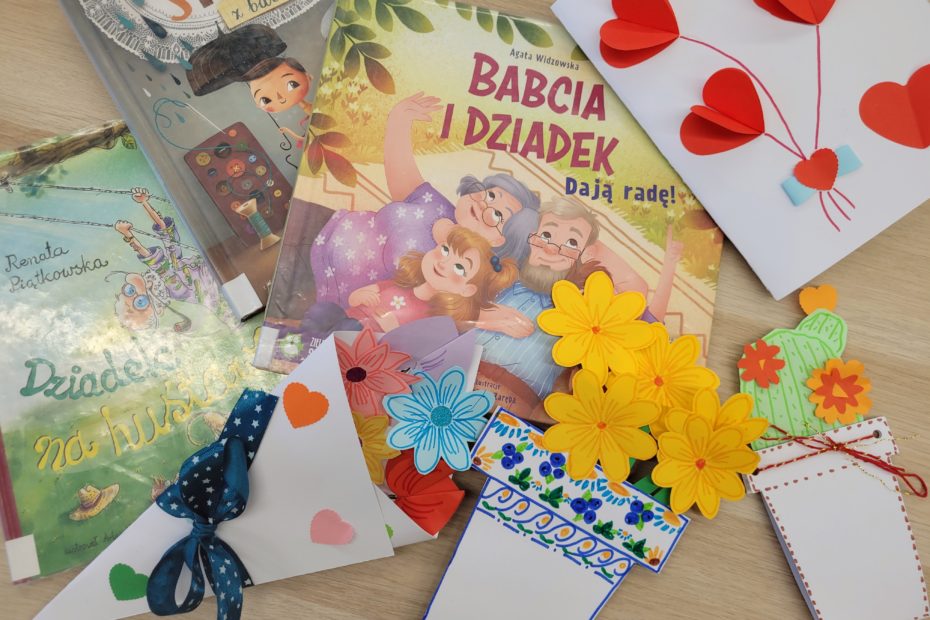 Na jasnym drewnianym stole leżą książki dla dzieci o babci i dziadku oraz kolorowe, ręcznie wykonane kartki i dekoracje: czerwone papierowe serca, żółte kwiaty, ozdobne zakładki i laurki. Całość tworzy ciepłą, radosną kompozycję z okazji Dnia Babci i Dziadka.