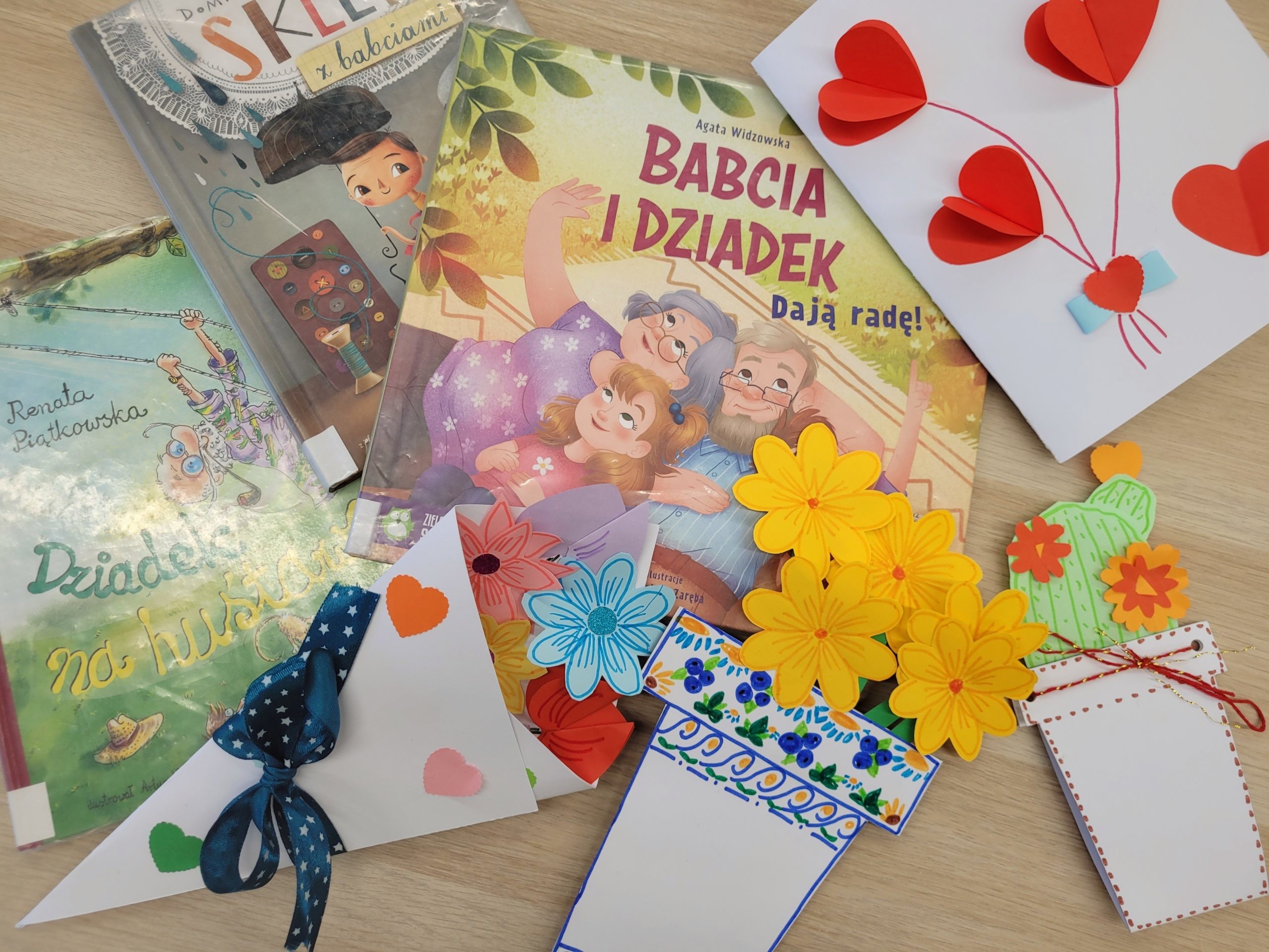 Na jasnym drewnianym stole leżą książki dla dzieci o babci i dziadku oraz kolorowe, ręcznie wykonane kartki i dekoracje: czerwone papierowe serca, żółte kwiaty, ozdobne zakładki i laurki. Całość tworzy ciepłą, radosną kompozycję z okazji Dnia Babci i Dziadka.