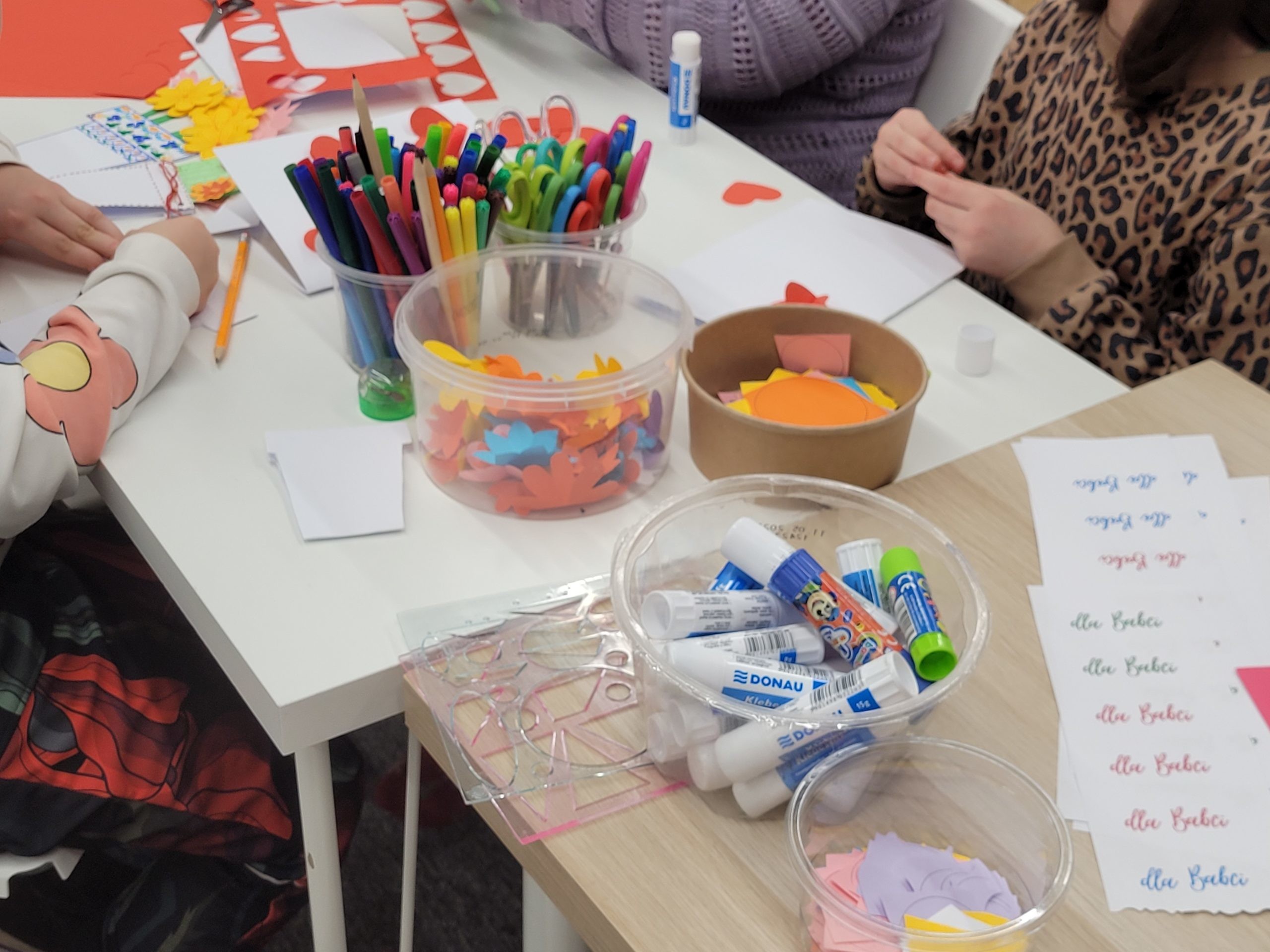 Warsztaty plastyczne w bibliotece. Dzieci wykonują papierowe kartki. Na stole pojemniki z klejami, nożyczkami, flamastrami i kolorowymi papierowymi elementami oraz naklejki z napisami "dla Babci", "dla Dziadka".