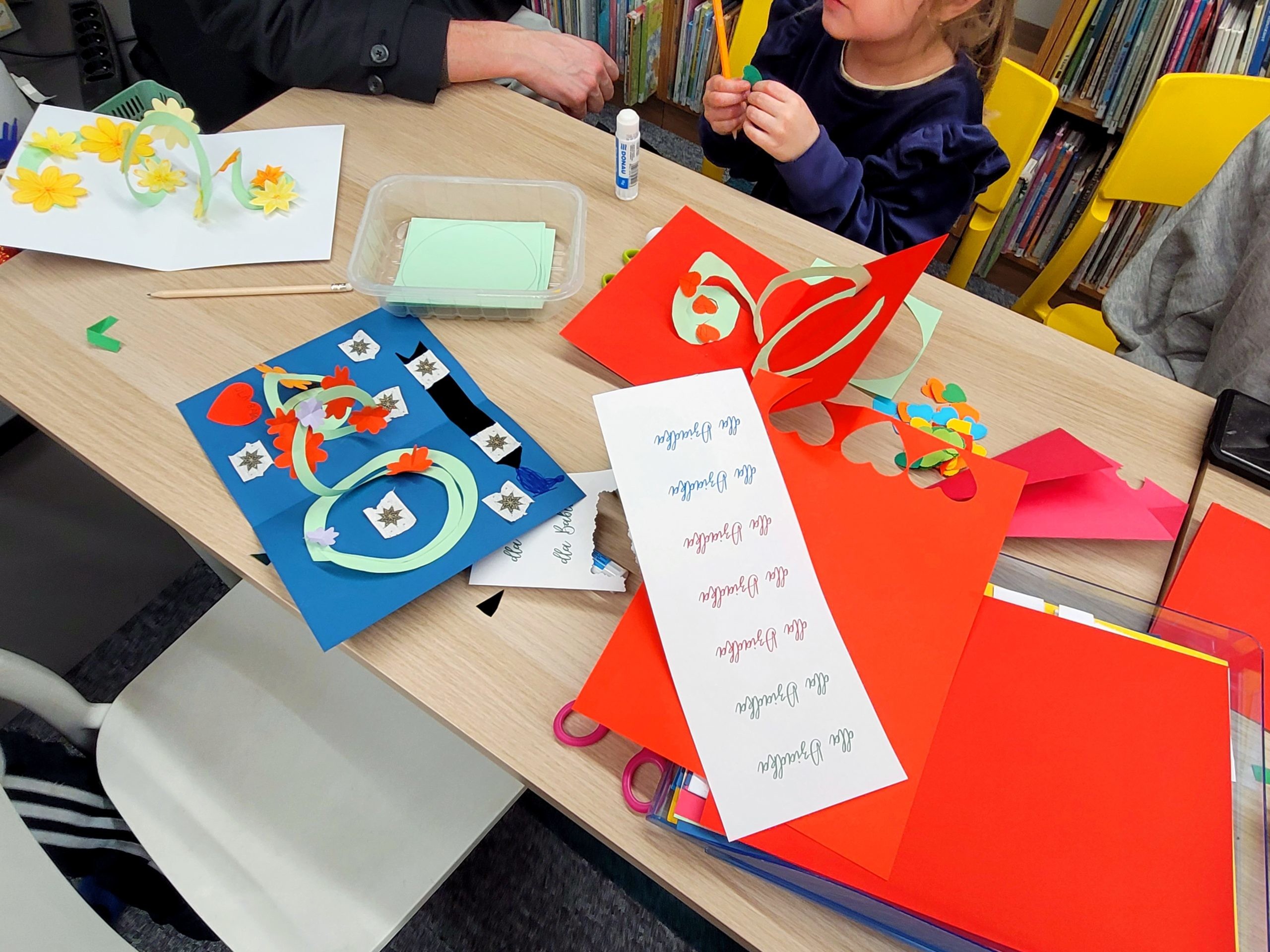 Warsztaty plastyczne w bibliotece. Dzieci i dorośli wykonują papierowe kartki z elementami 3d w środku. Na stole kartki, kolorowy papier oraz naklejki z napisami "dla Dziadka".