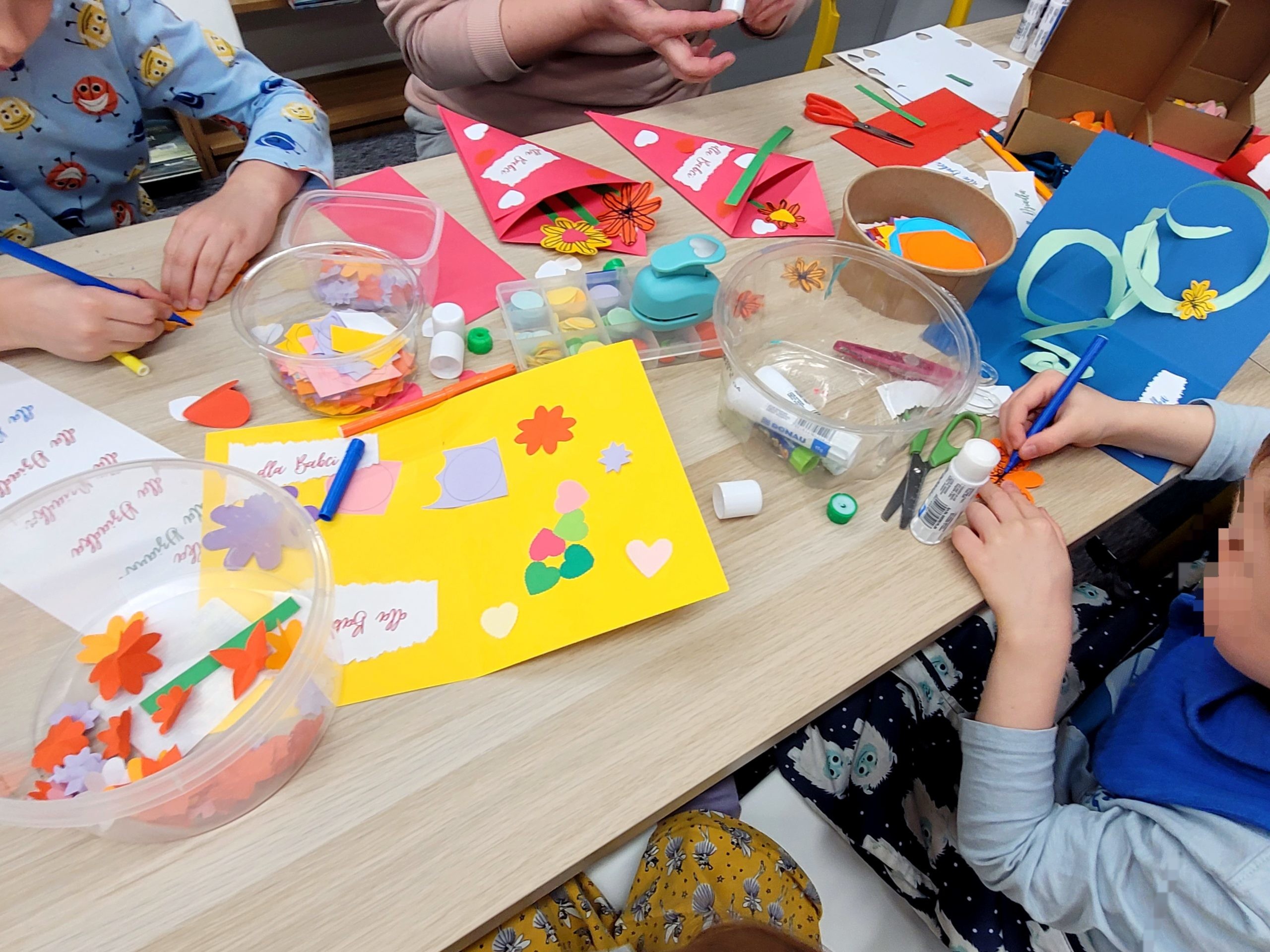Warsztaty plastyczne w bibliotece. Dzieci i dorośli wykonują papierowe kartki z elementami 3d w środku oraz papierowe bukiety. Na stole kartki, kolorowy papier oraz naklejki z napisami "dla Dziadka", "dla Babci".