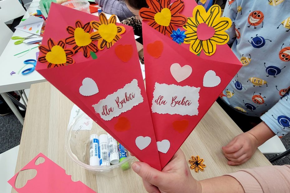 Ręka trzyma duże czerwone papierowe stożki z kwiatami (żółte i pomarańczowe), napisy "Dla Babci" i "Dla Babcia"; na stole klej i materiały plastyczne. W tle dzieci biorące udział w warsztatach plastycznych.