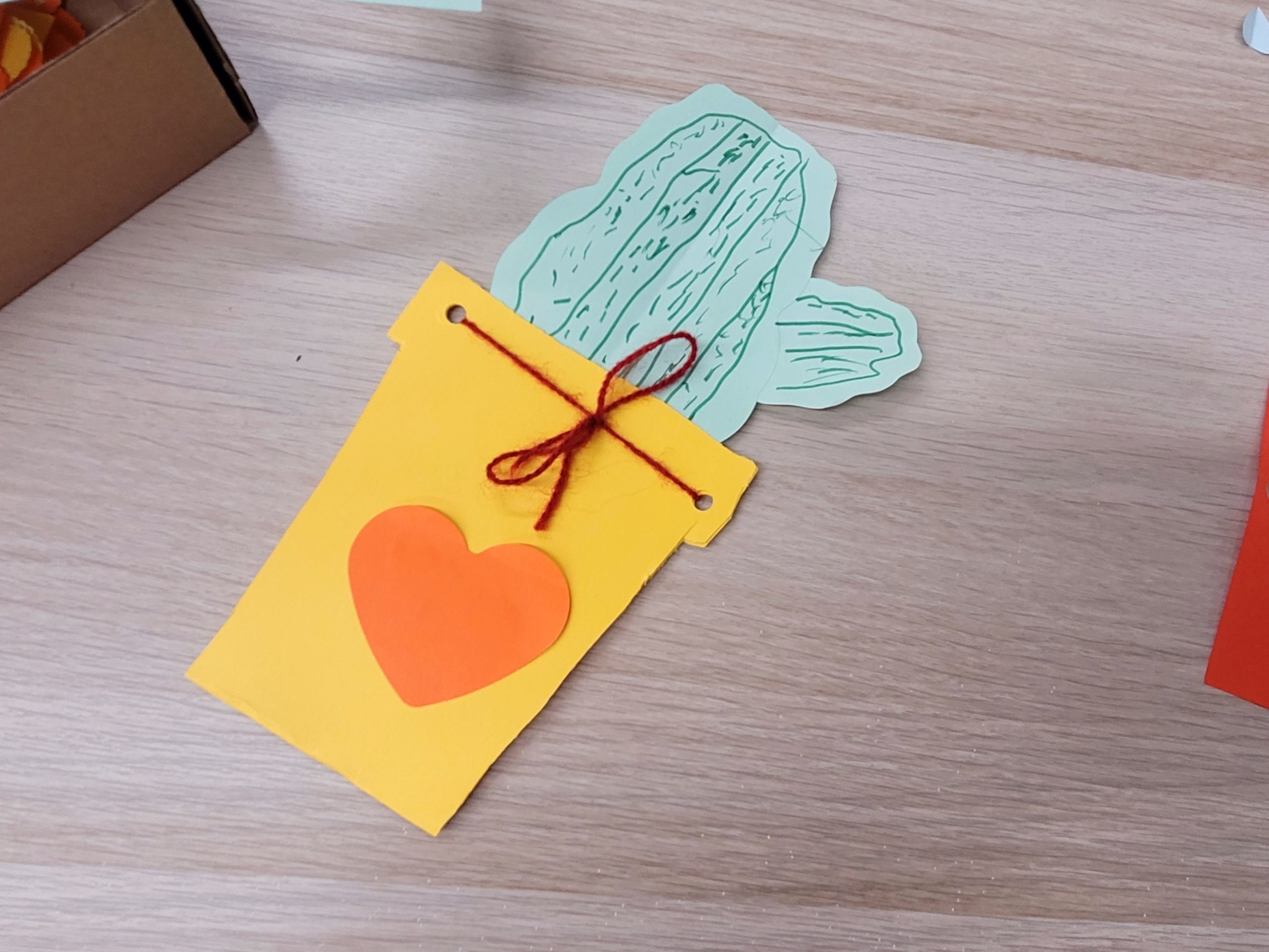 Żółta papierowa doniczka z zielonym kaktusem, czerwonym sercem i sznurkiem; obok pudełko i czerwony papier.
