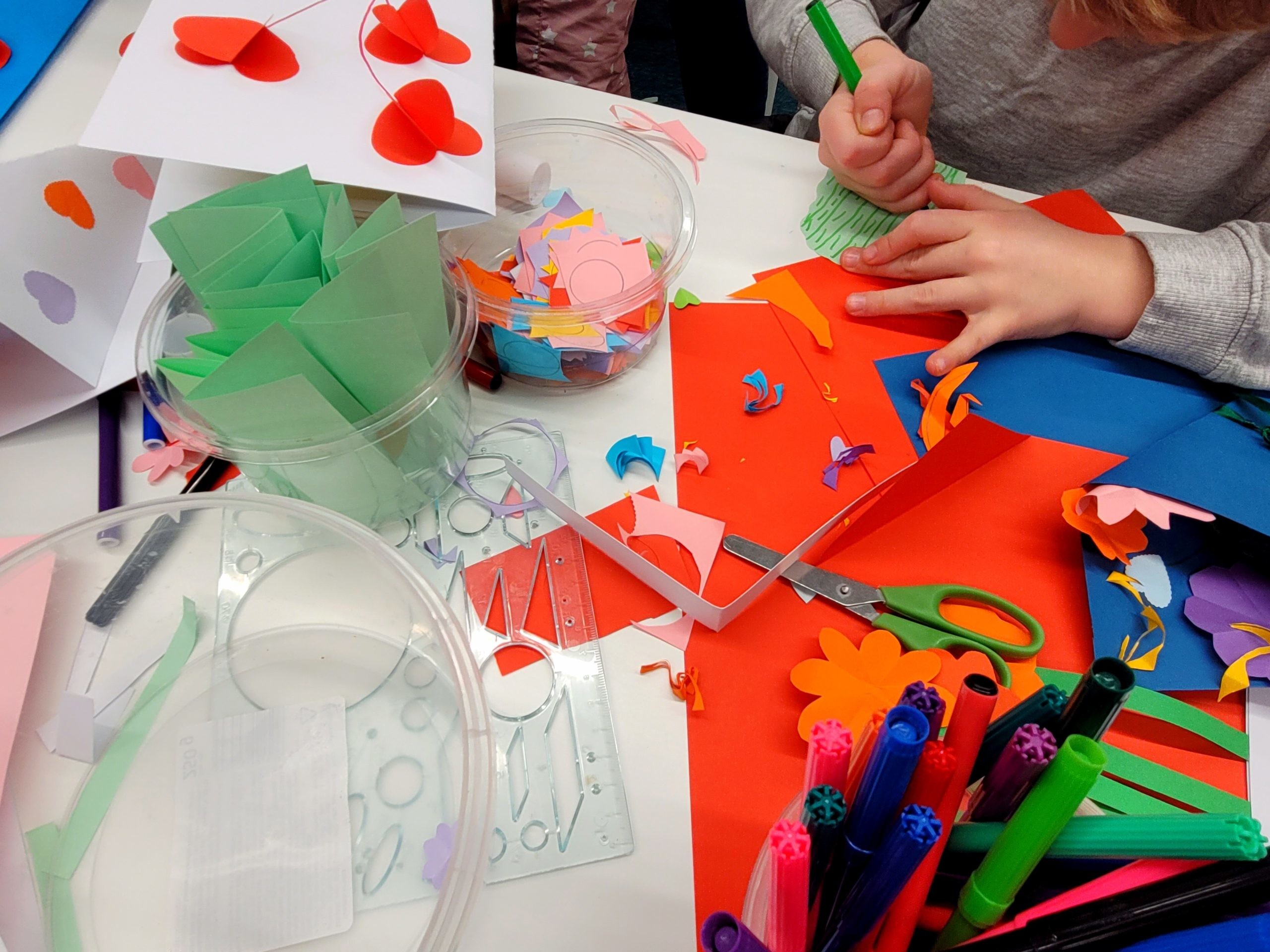 Warsztaty plastyczne w bibliotece. Dzieci wykonują papierowe kartki z elementami 3d w środku oraz papierowe bukiety. Na stole kartki, kolorowy papier, pojemniczki z papierowymi elementami, flamastry, nożyczki.