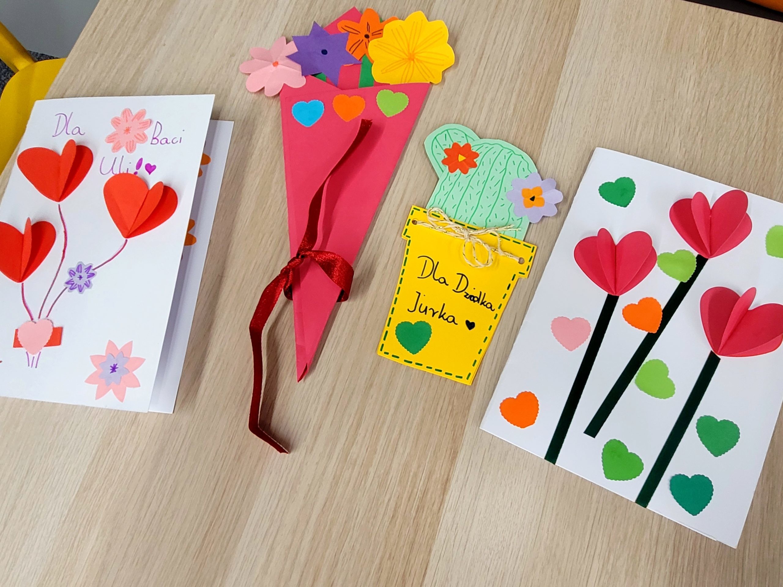 Ręcznie wykonane kartki na drewnianym stole: po lewej czerwona z sercami i kwiatami "Dla Babci", pośrodku stożkowaty bukiet z kolorowych kwiatów papierowych , po prawej żółty doniczkowy kaktus z sercem "Dla Dziadka", biała kartka z kolorowymi sercami.