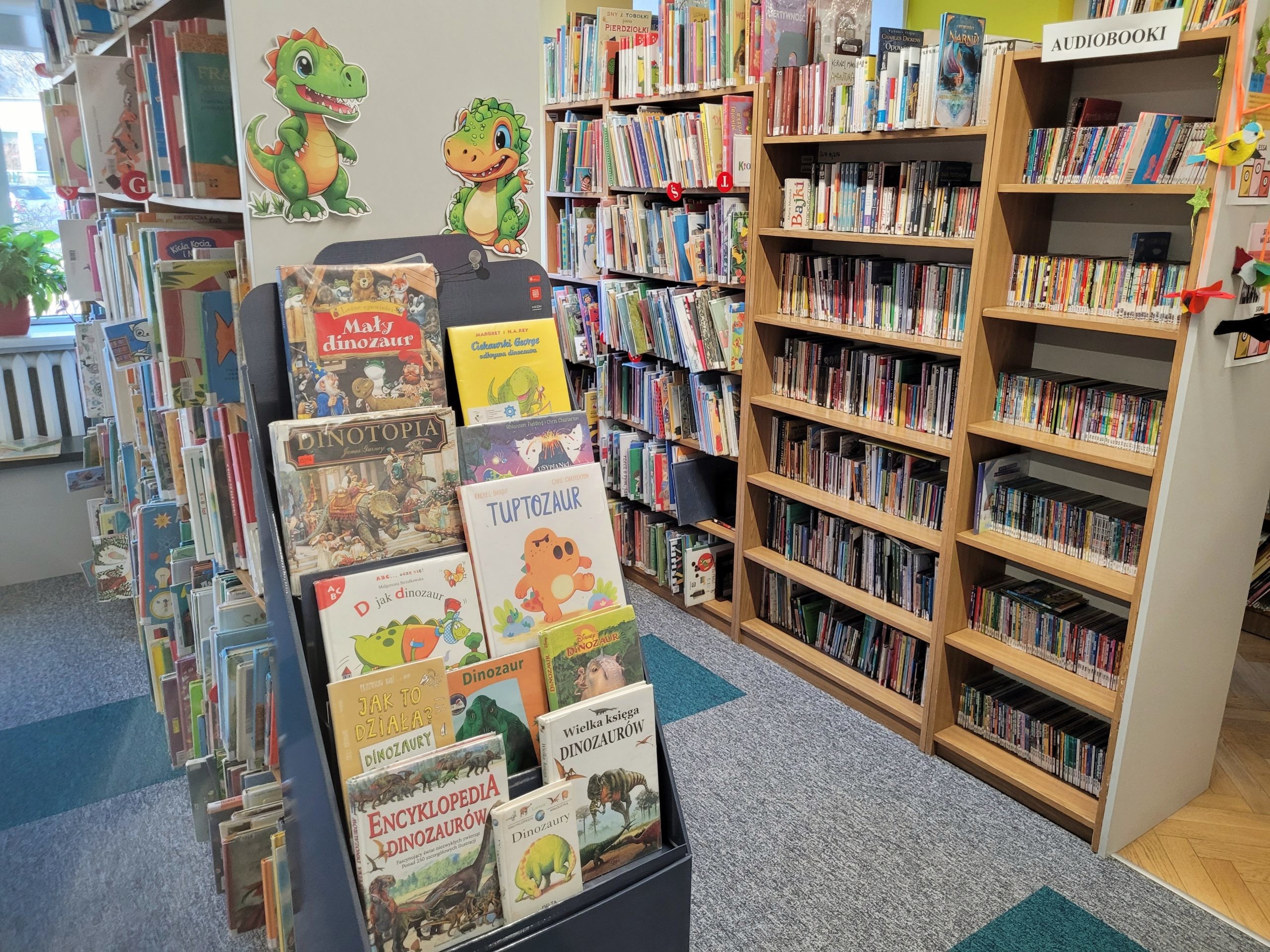 Wnętrze działu dziecięcego w bibliotece z drewnianymi regałami pełnymi książek. Na pierwszym planie znajduje się stojak z wyeksponowanymi książkami o dinozaurach, a nad nim kolorowe ilustracje uśmiechniętych dinozaurów przyklejone do ściany. Po prawej stronie widać półki z audiobookami, a całość tworzy przyjazną, uporządkowaną przestrzeń dla młodych czytelników.