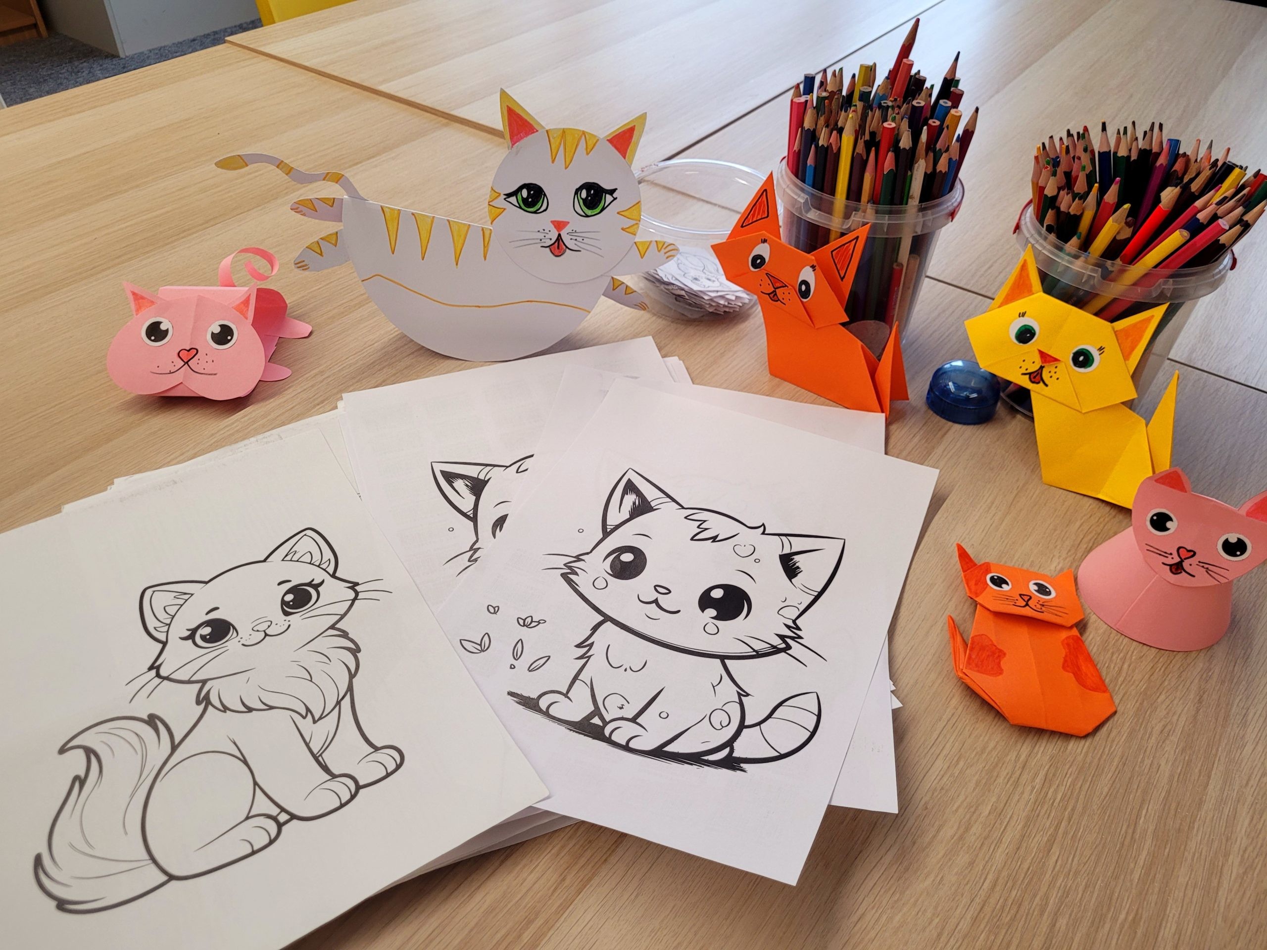 Stół z materiałami plastycznymi przygotowanymi na zajęcia z okazji Dnia Kota. Na blacie leżą kolorowanki z wizerunkami kotów, obok stoją pojemniki z kredkami oraz kolorowe papierowe koty wykonane techniką origami.