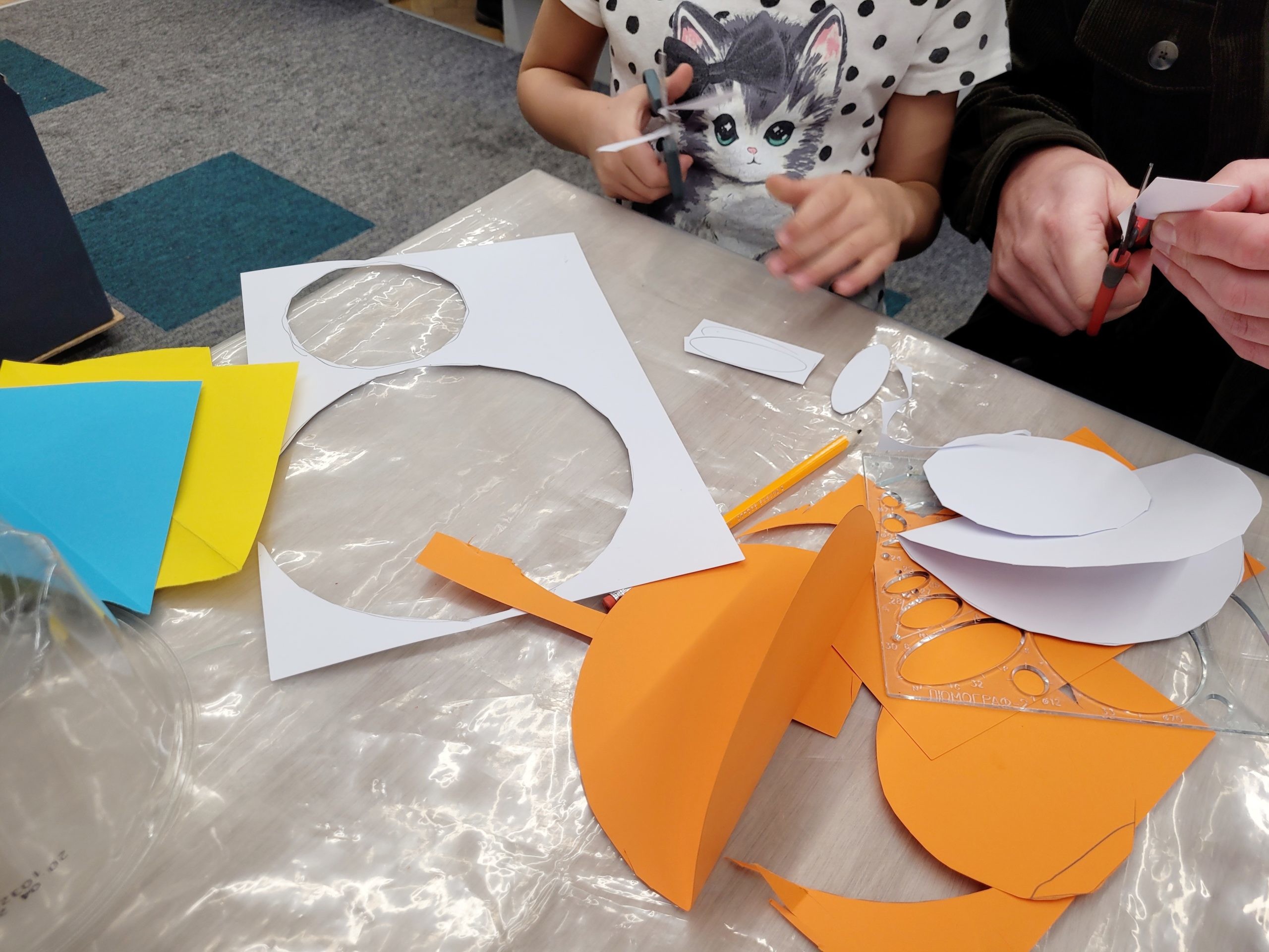 Dziecko i dorosły wycinają nożyczkami kształty z papieru przy stole warsztatowym. Na blacie leżą kolorowe kartki, ołówek i plastikowy szablon do rysowania figur.