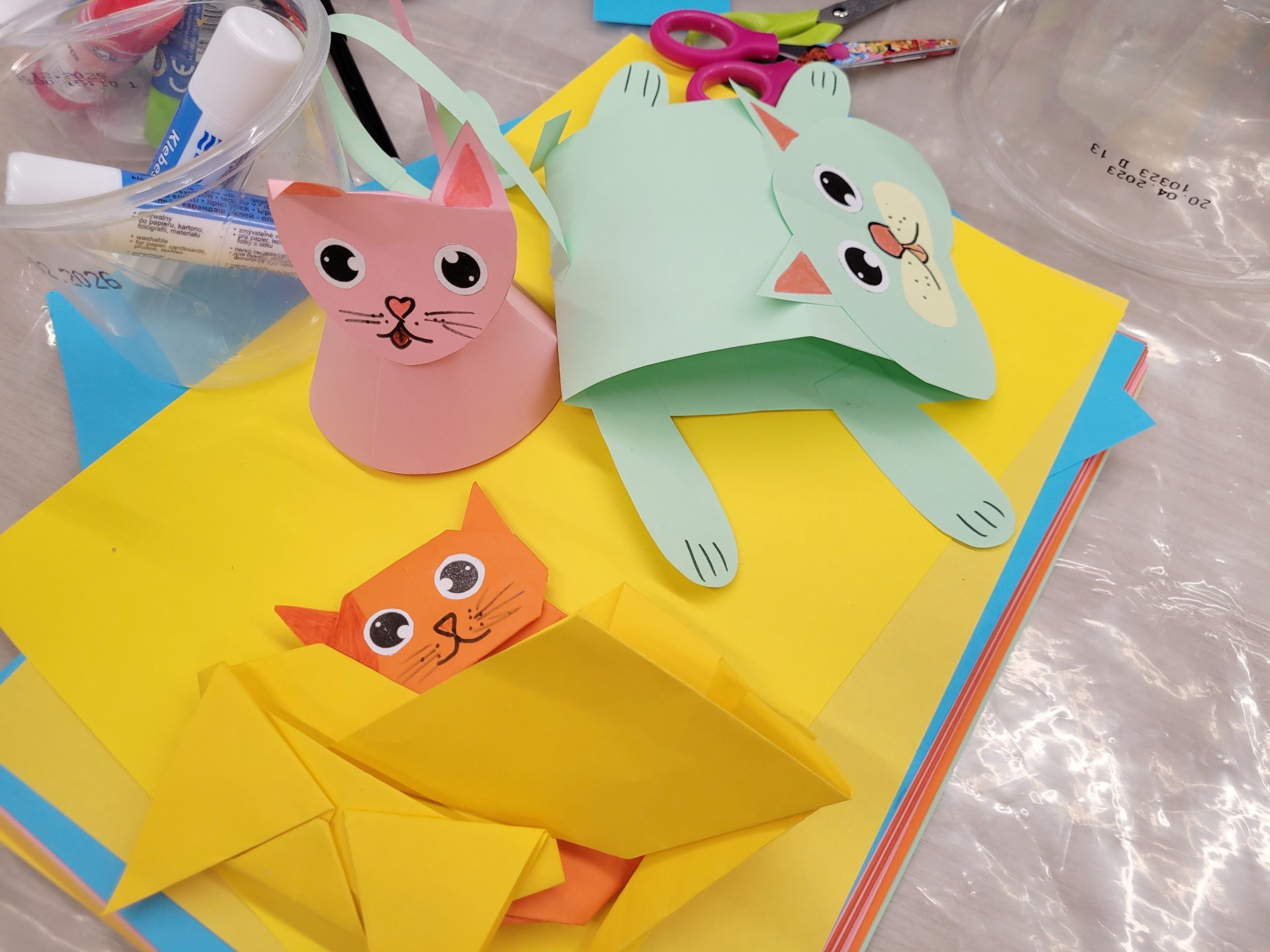 Kolorowe papierowe koty wykonane z wyciętych i złożonych kartek leżą na stole pokrytym żółtym papierem. Obok znajdują się nożyczki, klej w sztyfcie oraz plastikowe pojemniki z materiałami plastycznymi.