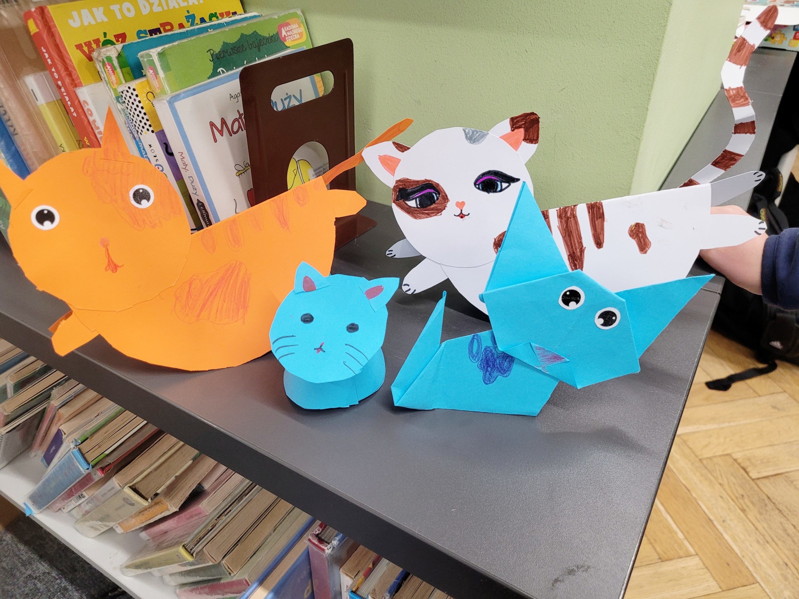 Kilka większych, kolorowych papierowych kotów stoi na półce obok książek. Wśród nich jest pomarańczowy kot, biały z brązowymi łatkami oraz dwa niebieskie.