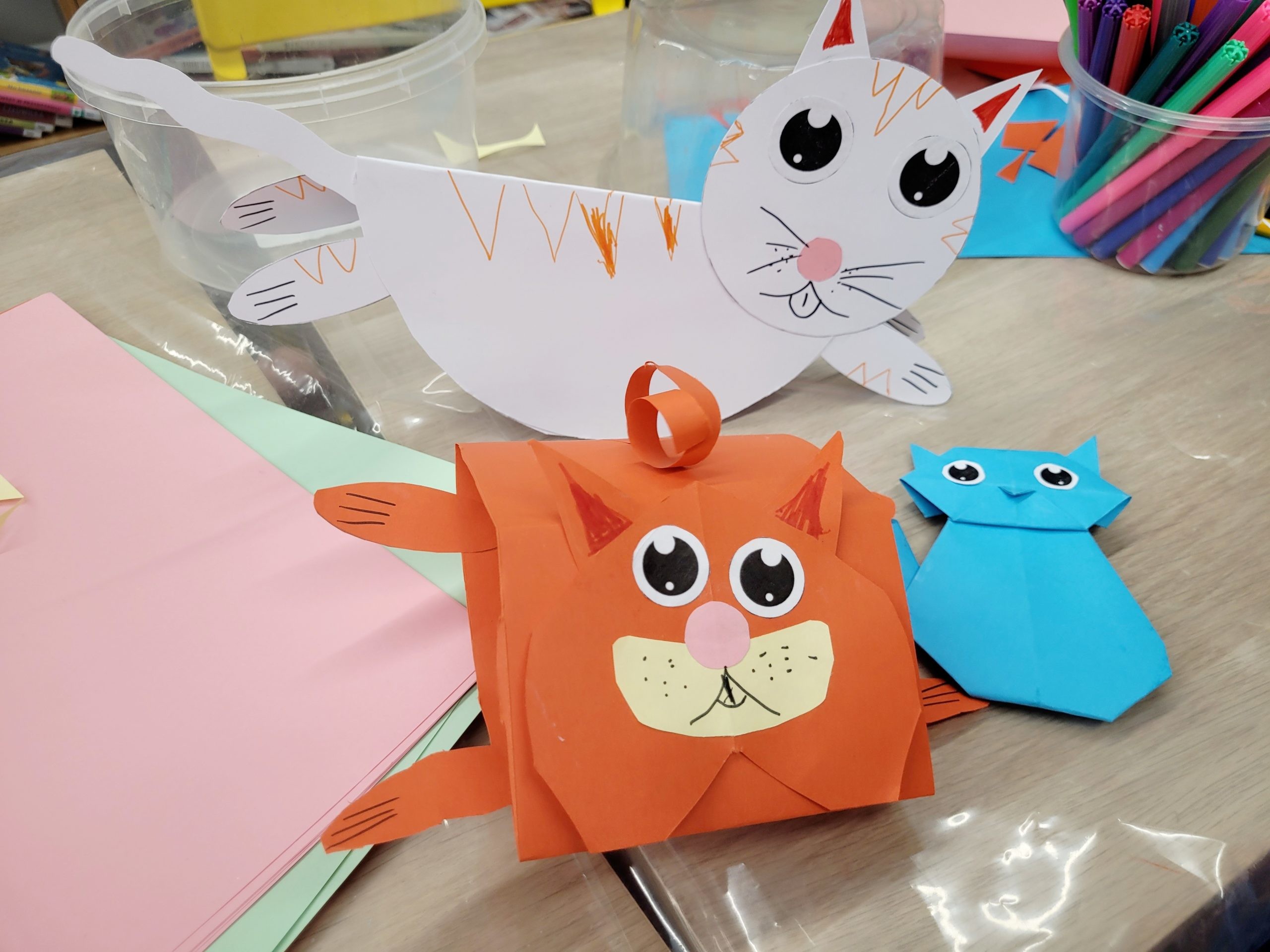 Kolorowe papierowe koty wykonane z wyciętych i złożonych kartek leżą na stole pokrytym żółtym papierem. Obok znajdują się flamastry, plastikowe pojemniki z materiałami plastycznymi oraz kolorowe kartki.