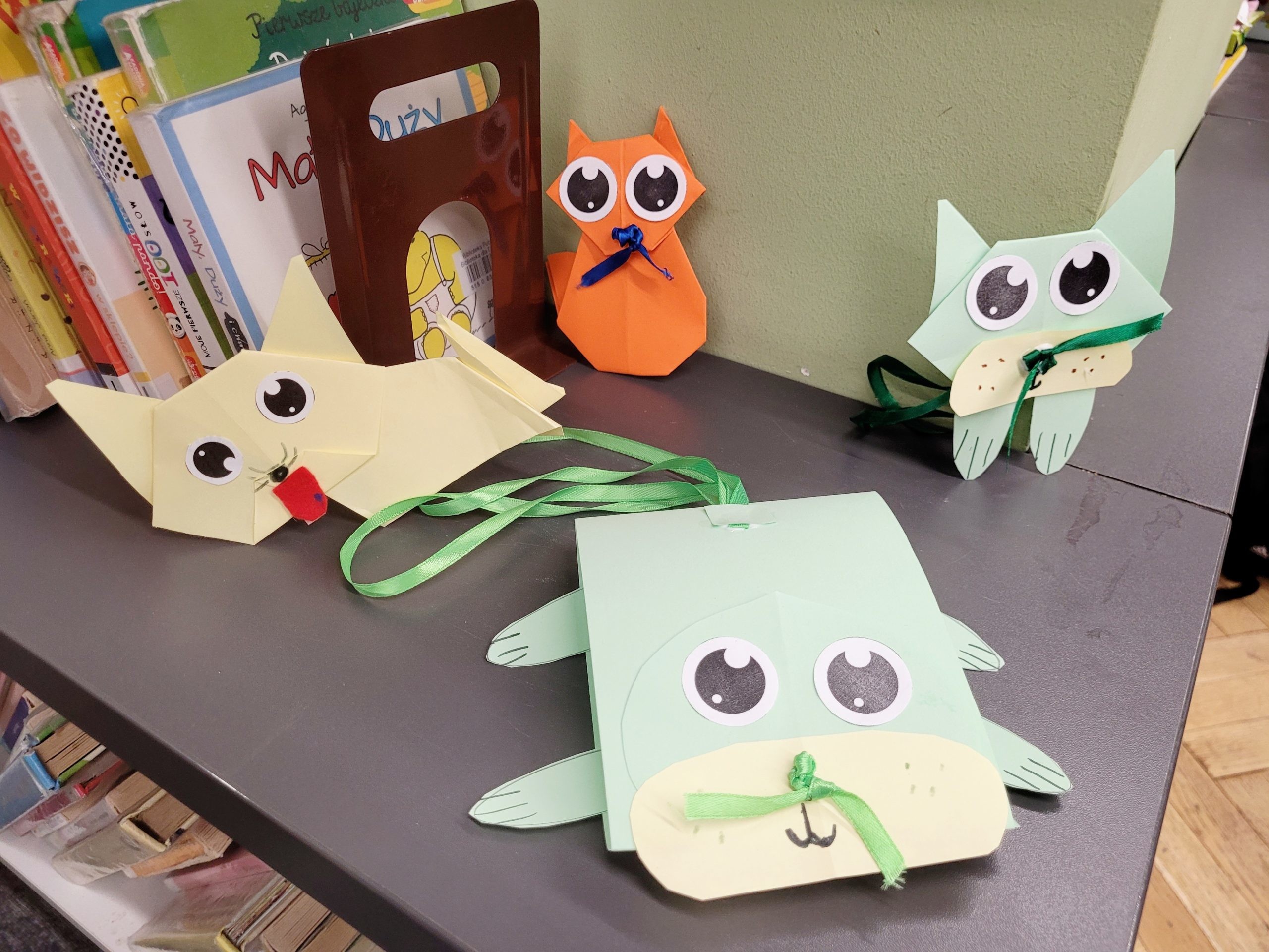 Kilka kolorowych papierowych kotów stoi na półce obok książek. Wśród nich jest pomarańczowy, dwa zielone i jeden jasnożółty.