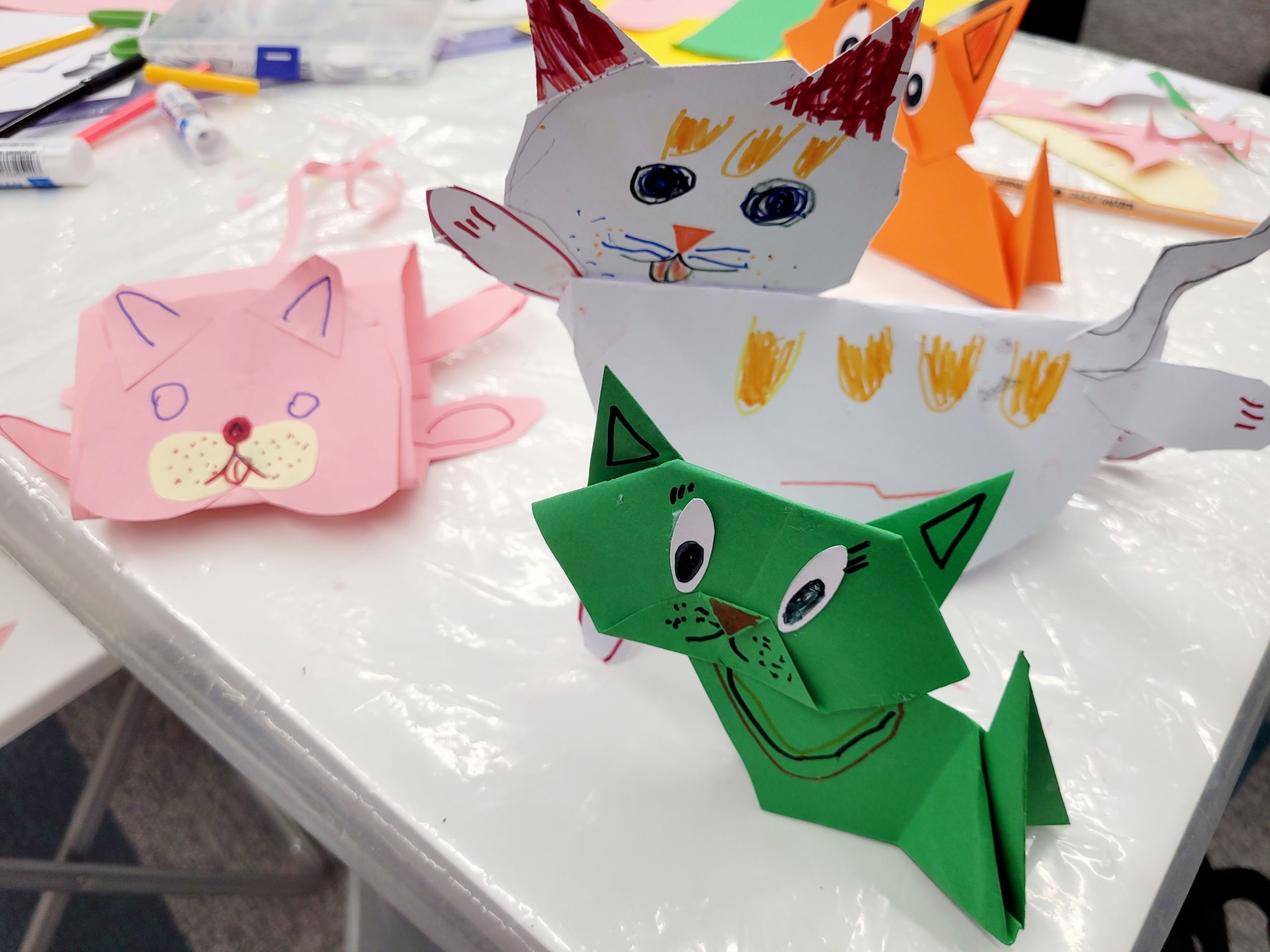 Cztery kolorowe papierowe koty wykonane z wyciętych i złożonych kartek leżą na stole pokrytym żółtym papierem. Obok znajdują się flamastry, kleje w sztyfcie oraz plastikowe pojemniki z materiałami plastycznymi oraz skrawki papieru.
