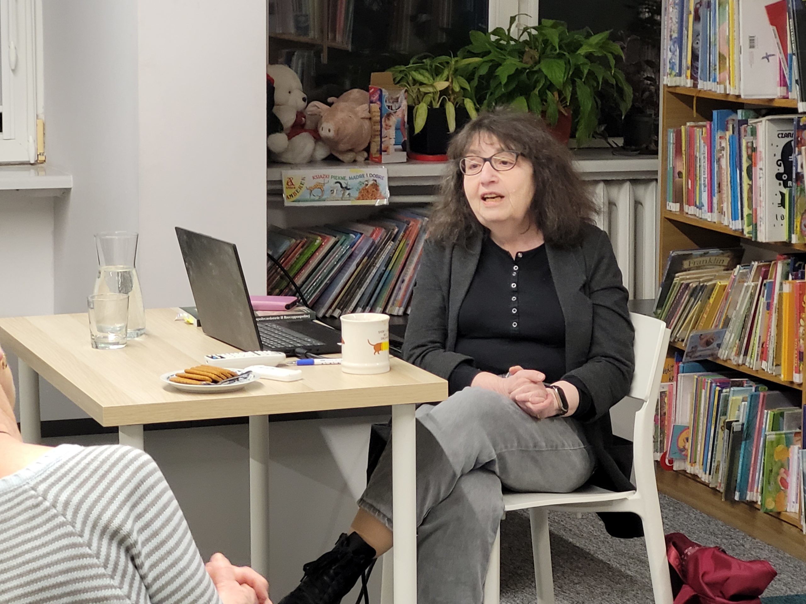 Kobieta siedzi przy stole w bibliotece i prowadzi spotkanie. Na stole stoi laptop, szklanka wody, kubek oraz talerzyk z ciastkami, a w tle widać półki z książkami i rośliny.