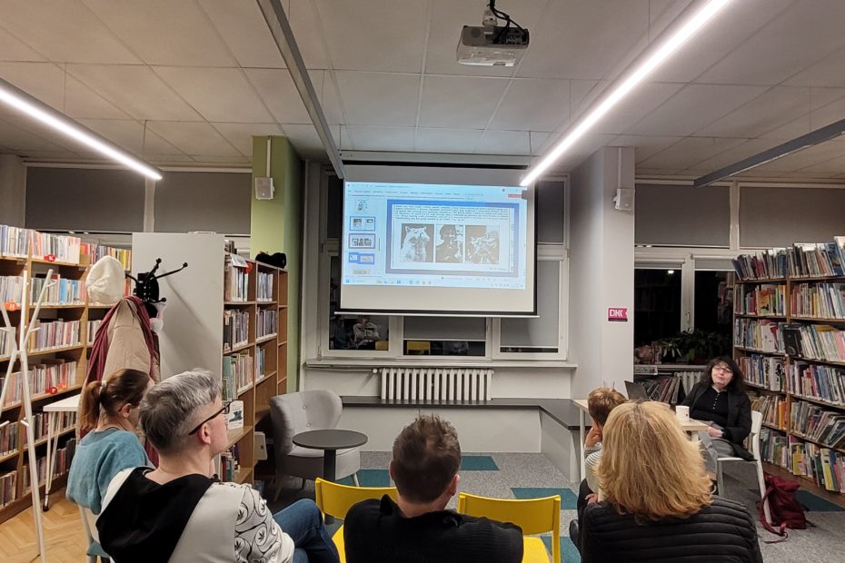 Grupa osób siedzi w bibliotece i słucha prezentacji wyświetlanej na ekranie. Na slajdzie widać reprodukcje trzech czarno-białych ilustracji lub fotografii umieszczonych obok siebie oraz fragment tekstu nad nimi. Prelegentka siedzi z boku, a wokół znajdują się regały z książkami i rzutnik pod sufitem.