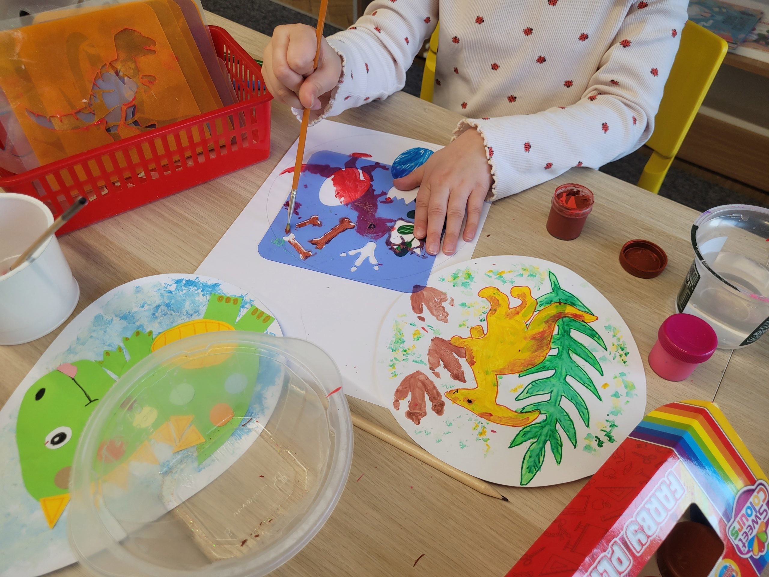 Dziecko maluje farbami obraz z dinozaurem przy stoliku. Na stole leżą kolorowe prace plastyczne, pędzel, kubek z wodą i wycinanki w kształcie dinozaurów.