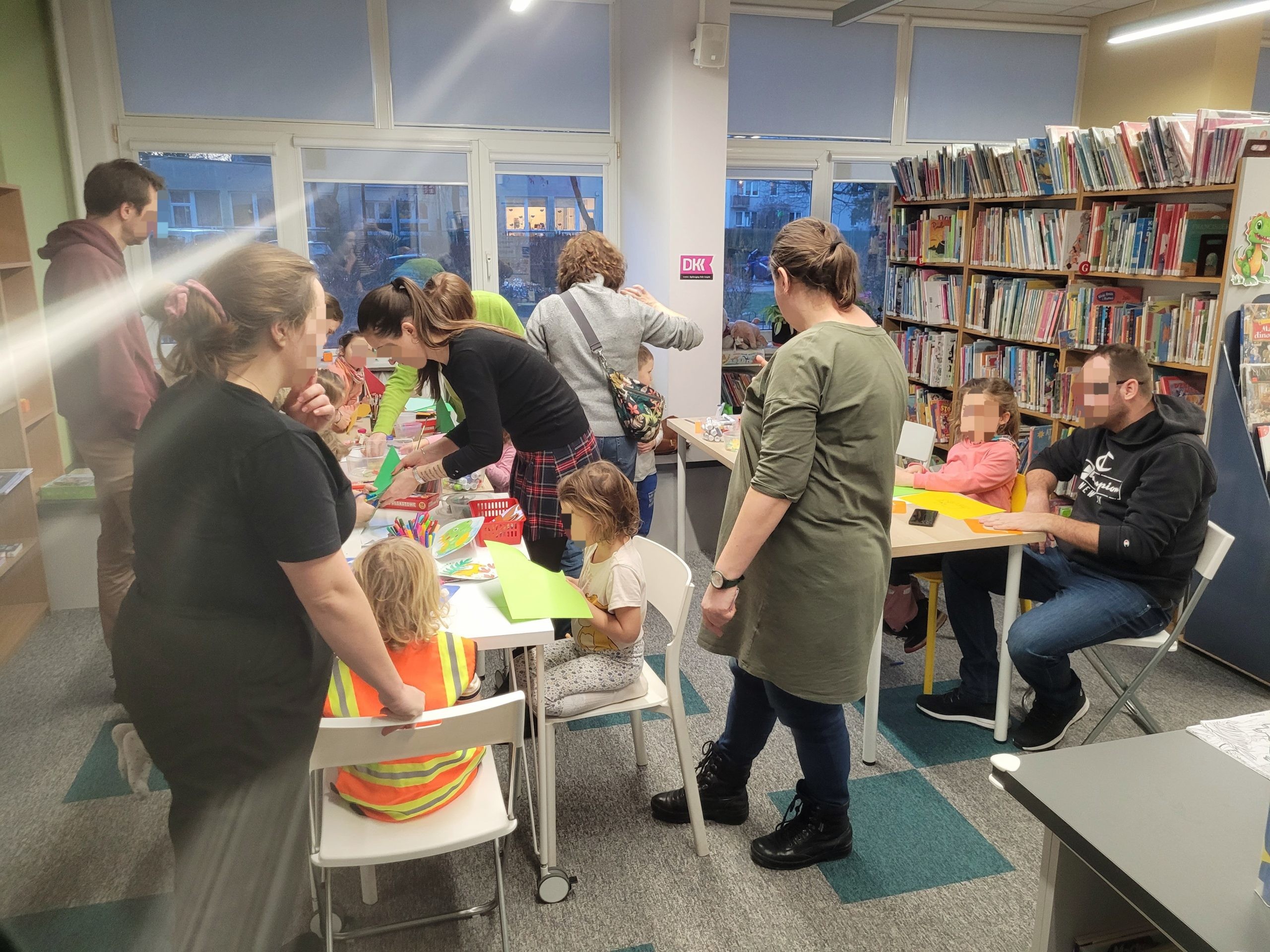 Grupa dzieci wraz z opiekunami uczestniczy w warsztatach plastycznych w bibliotece. Uczestnicy stoją i siedzą przy stołach, wycinając i przyklejając kolorowy papier. Na stołach leżą kartki, nożyczki, klej i flamastry. W tle widać wysokie regały wypełnione książkami dla dzieci oraz duże okna, przez które wpada światło dzienne.