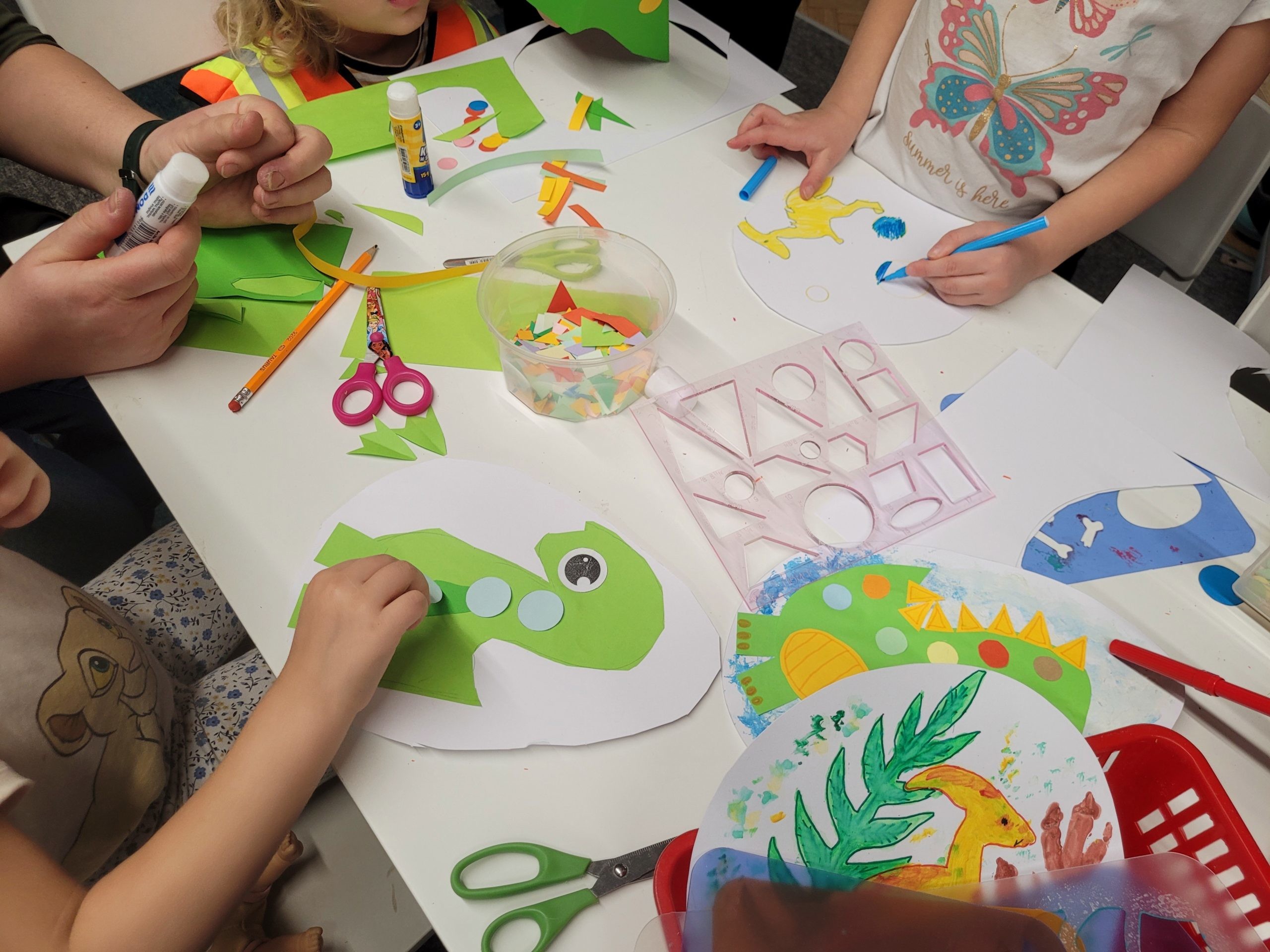 Zbliżenie na stół, przy którym dzieci tworzą prace plastyczne z motywem dinozaurów. Na białych kartkach widoczne są kolorowe sylwetki dinozaurów wyklejane z papieru oraz rysunki wykonane kredkami i flamastrami. Wokół rozłożone są przybory: klej w sztyfcie, nożyczki, szablony figur geometrycznych i małe kawałki kolorowego papieru.