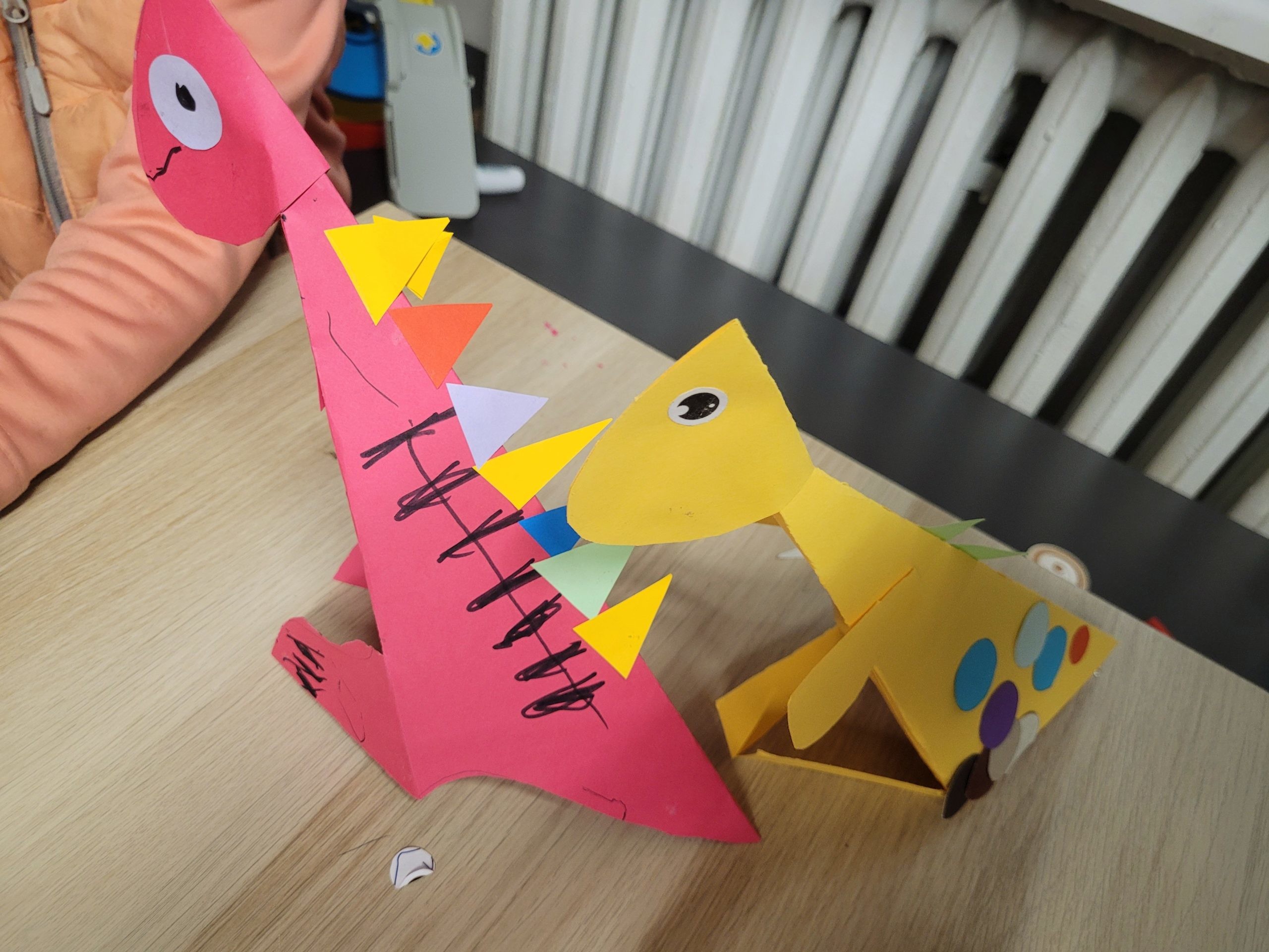 Różowy i żółty dinozaur z kolorowymi kółkami stoją na stole w bibliotece. W tle widać rękę dziecka oraz biały kaloryfer.