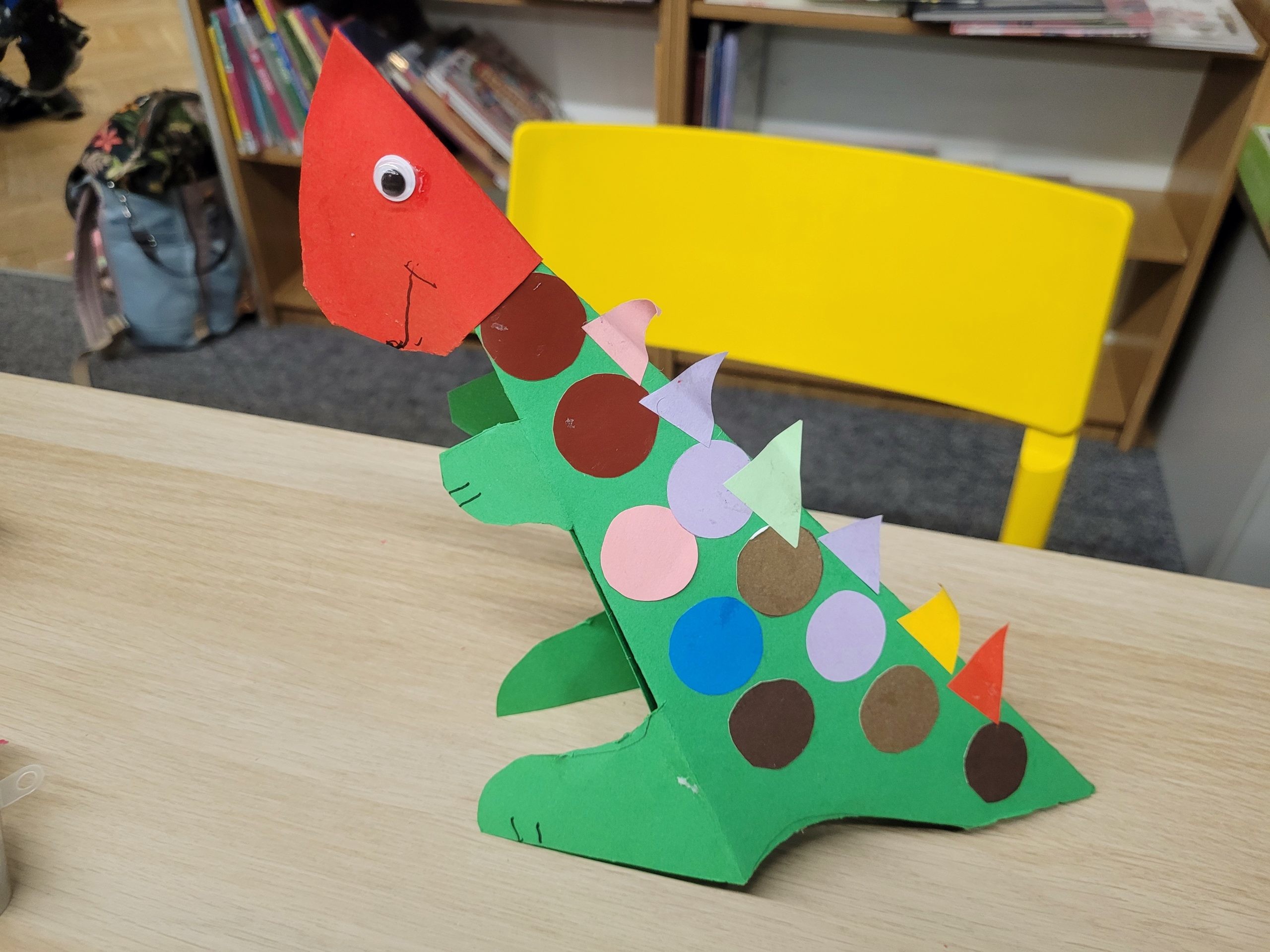 Papierowy zielony dinozaur z kolorowymi kółkami i ruchomym oczkiem stoi na stole w bibliotece. W tle widać regały z książkami.