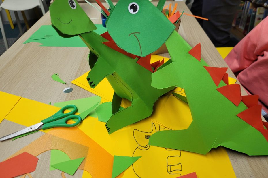 Dwa zielone dinozaury wykonane z papieru z czerwonymi trójkątami na grzbiecie stoją na stole pełnym wycinanek i nożyczek. W tle widać półki z książkami.