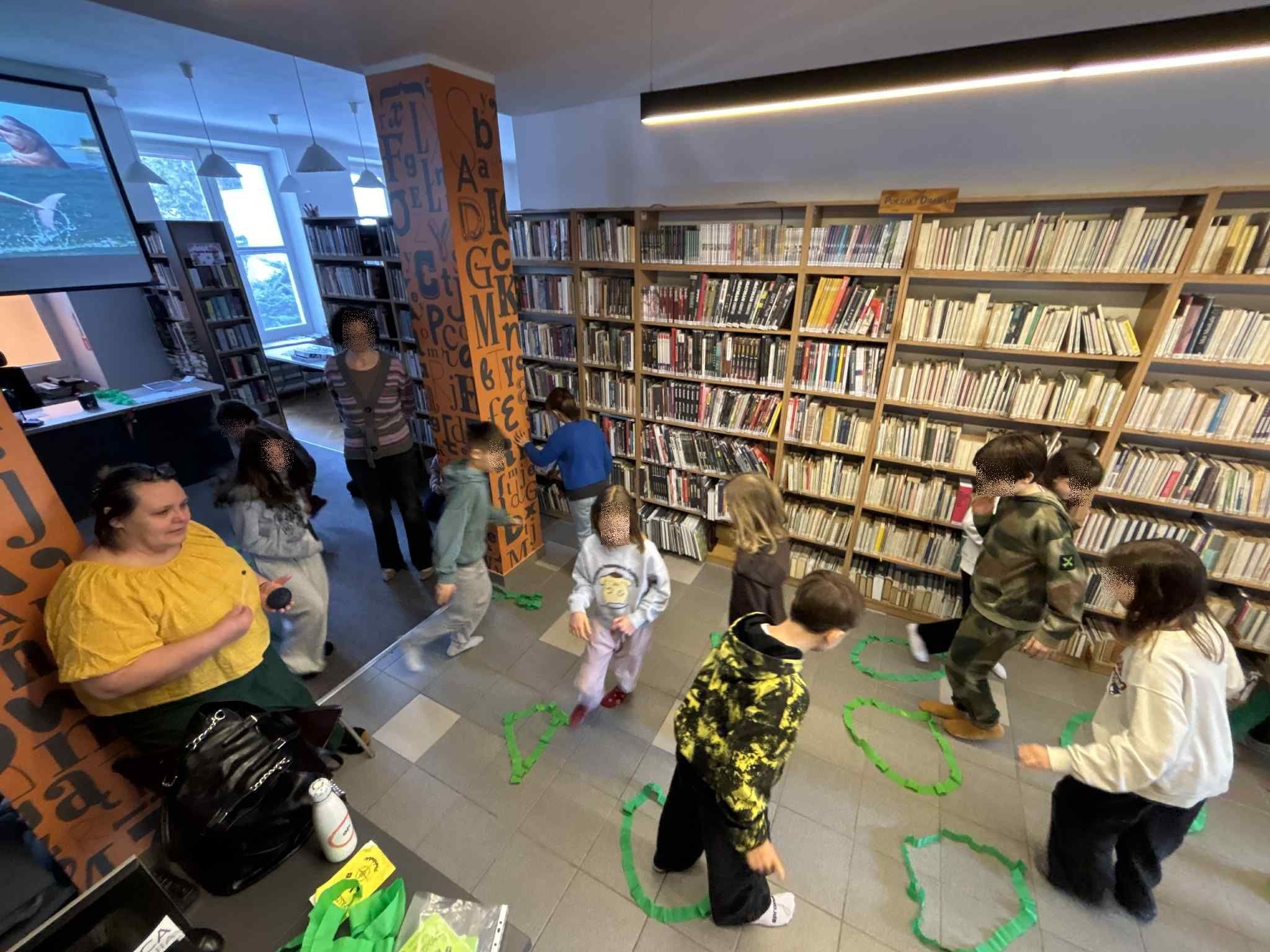 zdjęcie przedstawia zabawę ruchową dzieci. Na podłodze są koła zrobione z zielonej krepiny, dzieci biegają między nimi, po lewej stronie jest prowadząca, która trzyma w ręku mały instrument muzyczny.