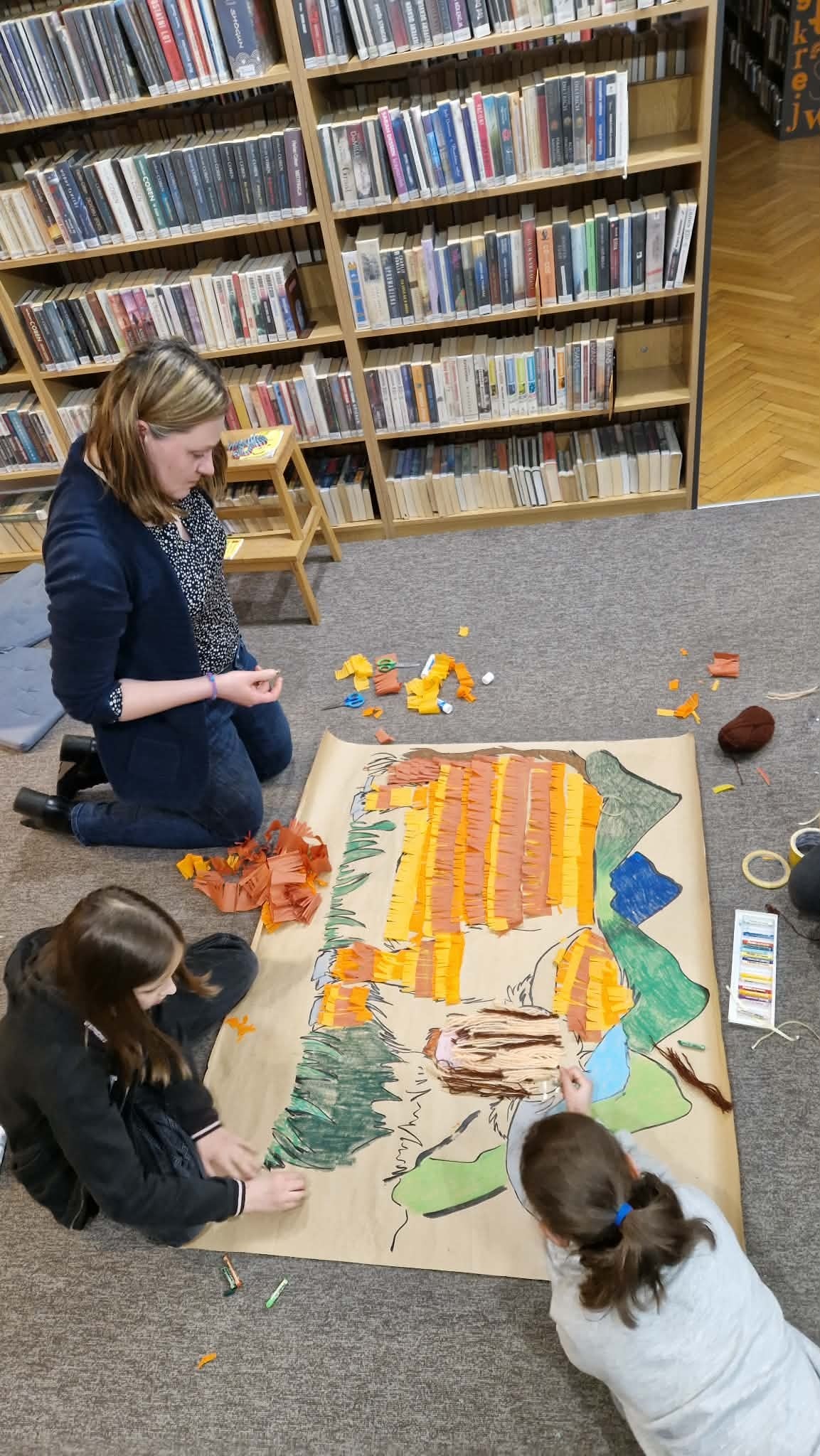 zjęcie przedstawia uczestników warsztatów podróżniczych. W centralnej części znajduje się rodzic, który pomaga w kończeniu pracy. U dołu zdjęcia są dwie pochylone dziewczynki .
