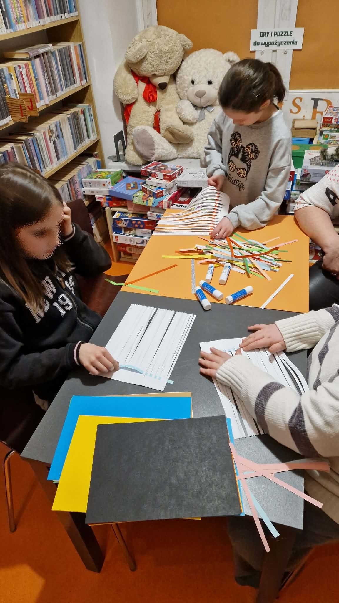 na zdjęciu widać uczestników zajęć. Siedzą przy stole i wykonują z papieru plecionkę, która ma przypominać szkocką kratę.