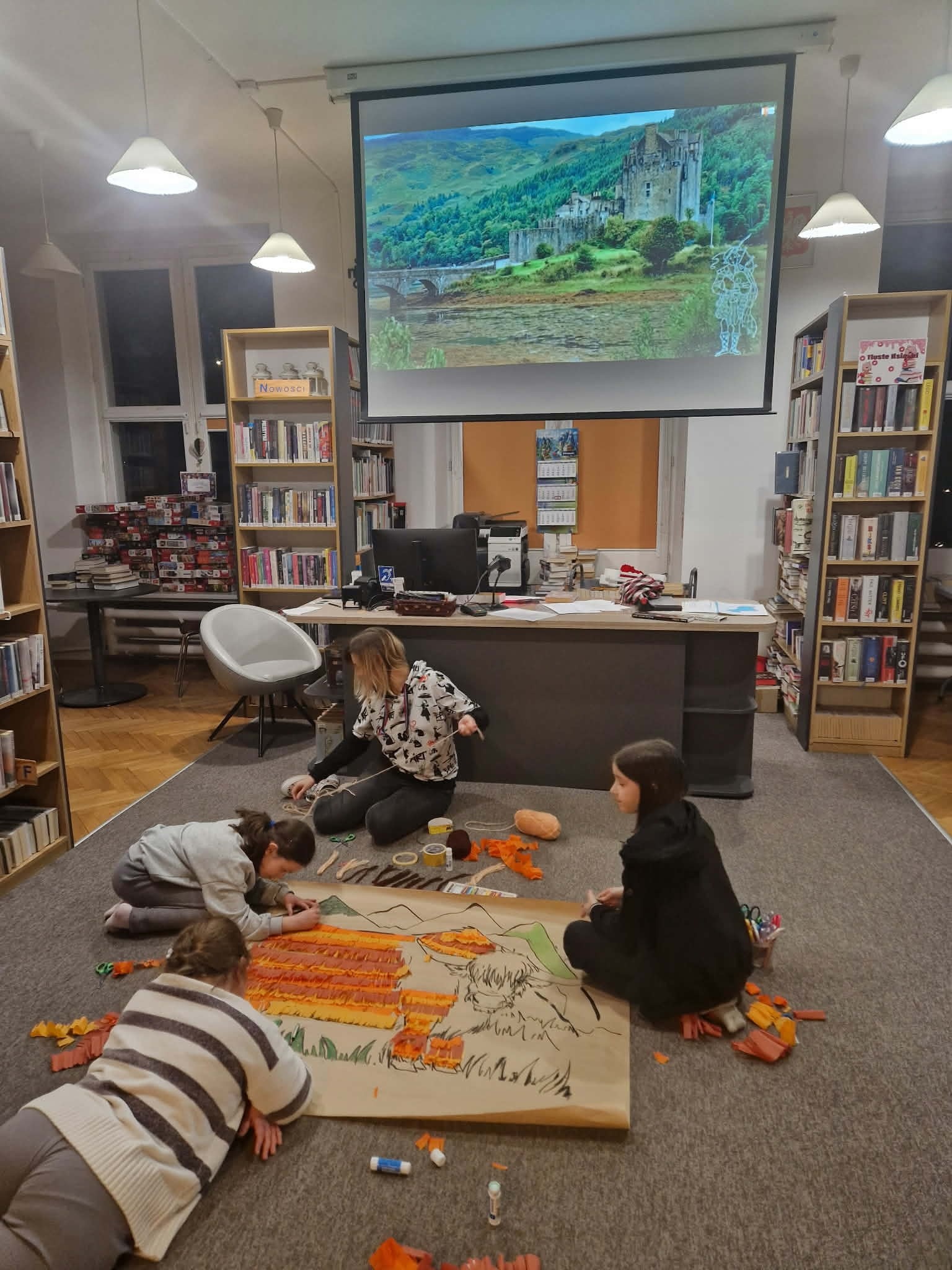 zdjęcie przedstawia dzieci podczas zajęć plastycznych związanych ze Szkocją. Trzy dziewczynki razem z prowadzącą pochylaja sie nad szarym papierem, na którym widnieje wizerunek krowy. Na papierze są paski kolorowych bibuł, nożyczki, kleje i włóczka.