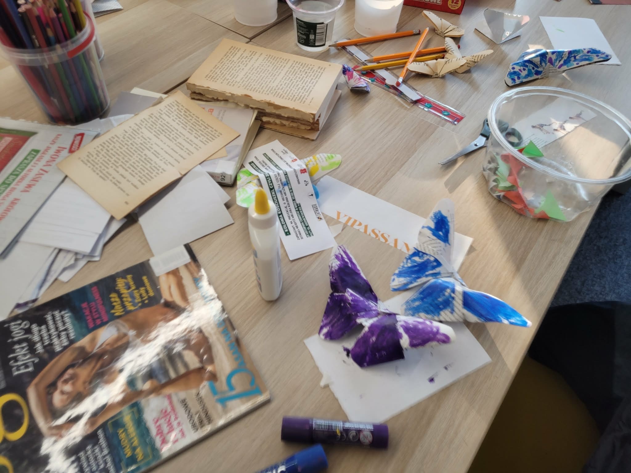 Stół warsztatowy w bibliotece z rozłożonymi materiałami plastycznymi: kolorowe kredki, klej, nożyczki, papier oraz wycinane i malowane papierowe motyle.