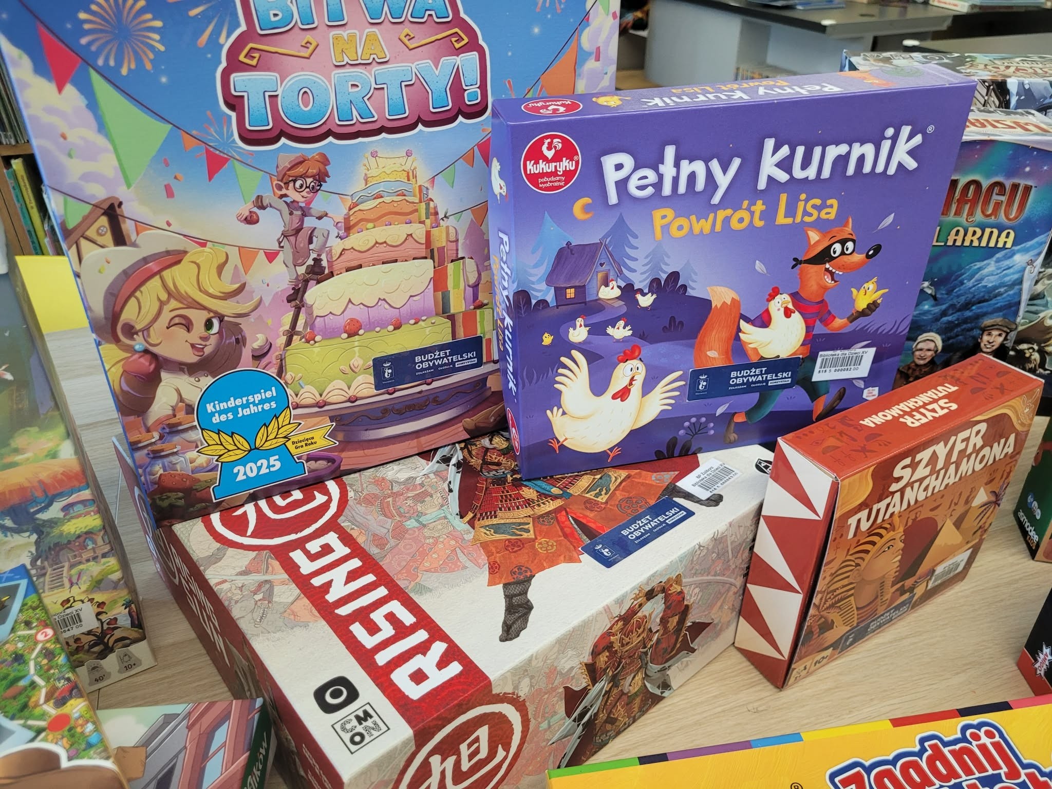 Różne gry planszowe dla dzieci i rodzin na stole w bibliotece, w tym „Bitwa na torty!”, „Pełny kurnik. Powrót lisa”, „Sabotażysta” i „Szyfr Tutanchamona”.