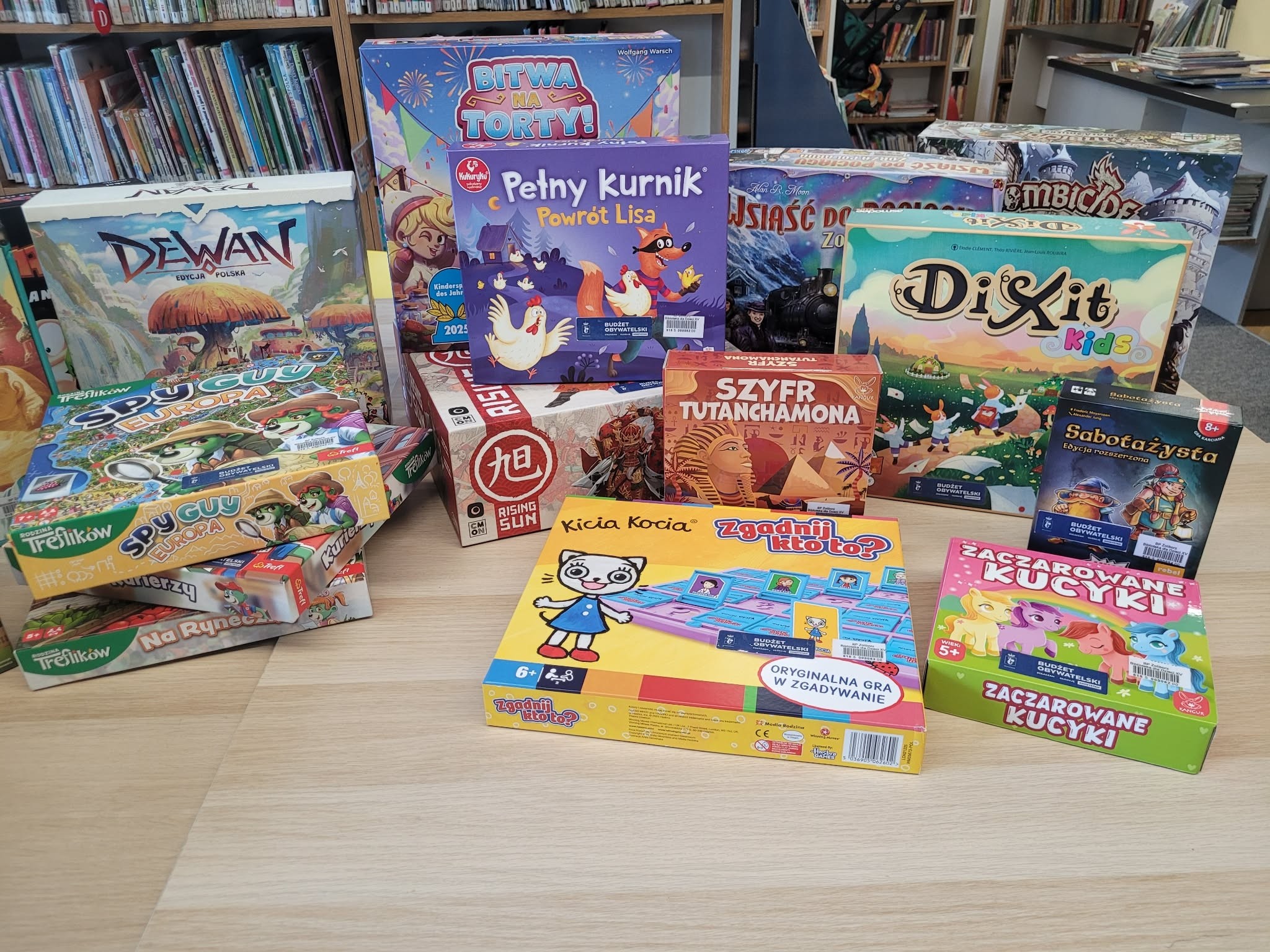 Na stole w bibliotece ustawione są gry planszowe z naklejką „Budżet Obywatelski”, m.in. „Dewan”, „Spy Guy Europa”, „Pełny Kurnik. Powrót Lisa”, „Bitwa na torty!”, „Dixit Kids”, „Szyfr Tutanchamona”, „Zgadnij kto to?”, „Sabotażysta” oraz „Zaczarowane kucyki”. W tle widoczne regały z książkami.