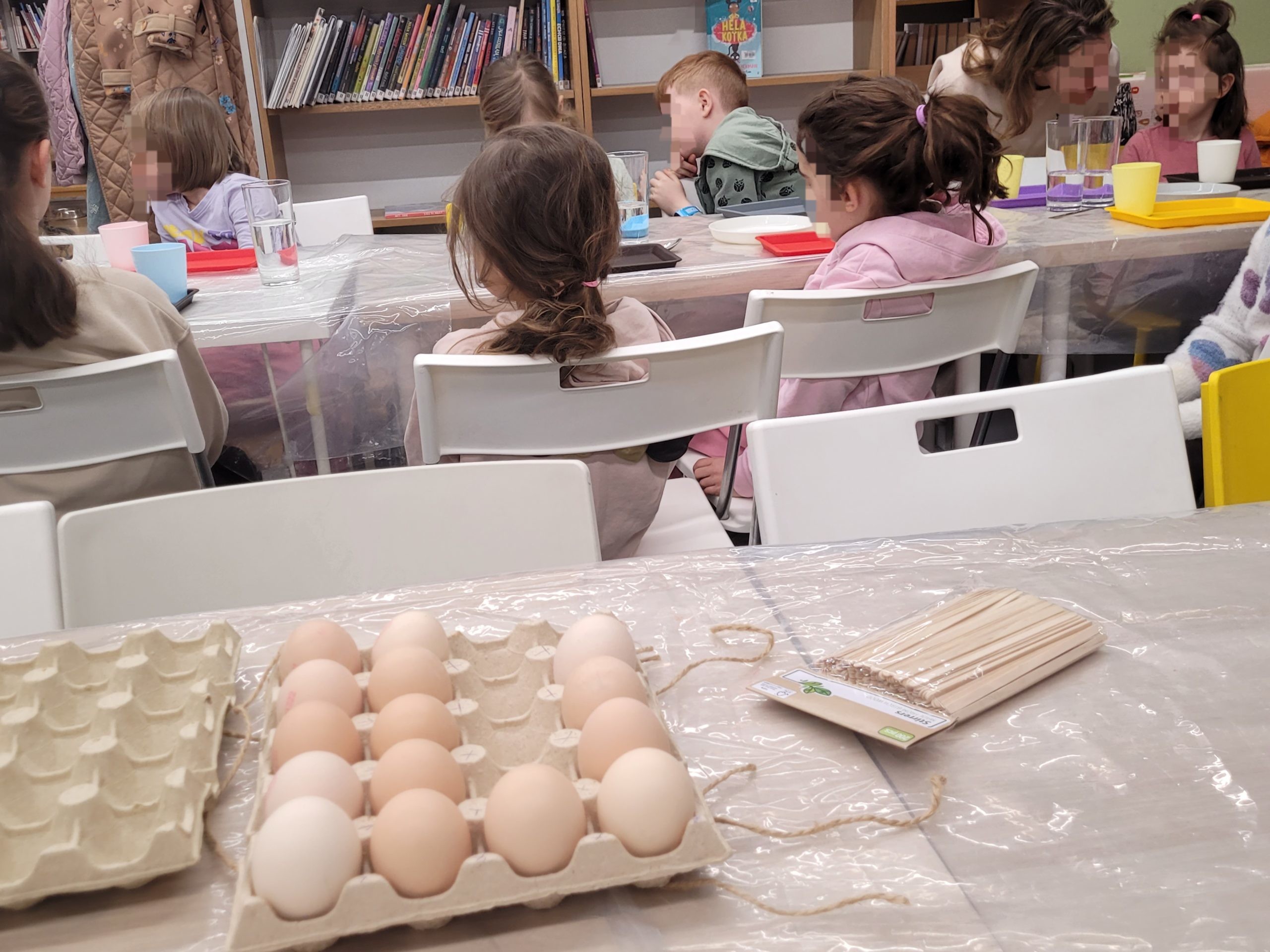 Na pierwszym planie tacki z jajkami i drewniane patyczki, w tle dzieci siedzące przy stole podczas warsztatów w bibliotece.