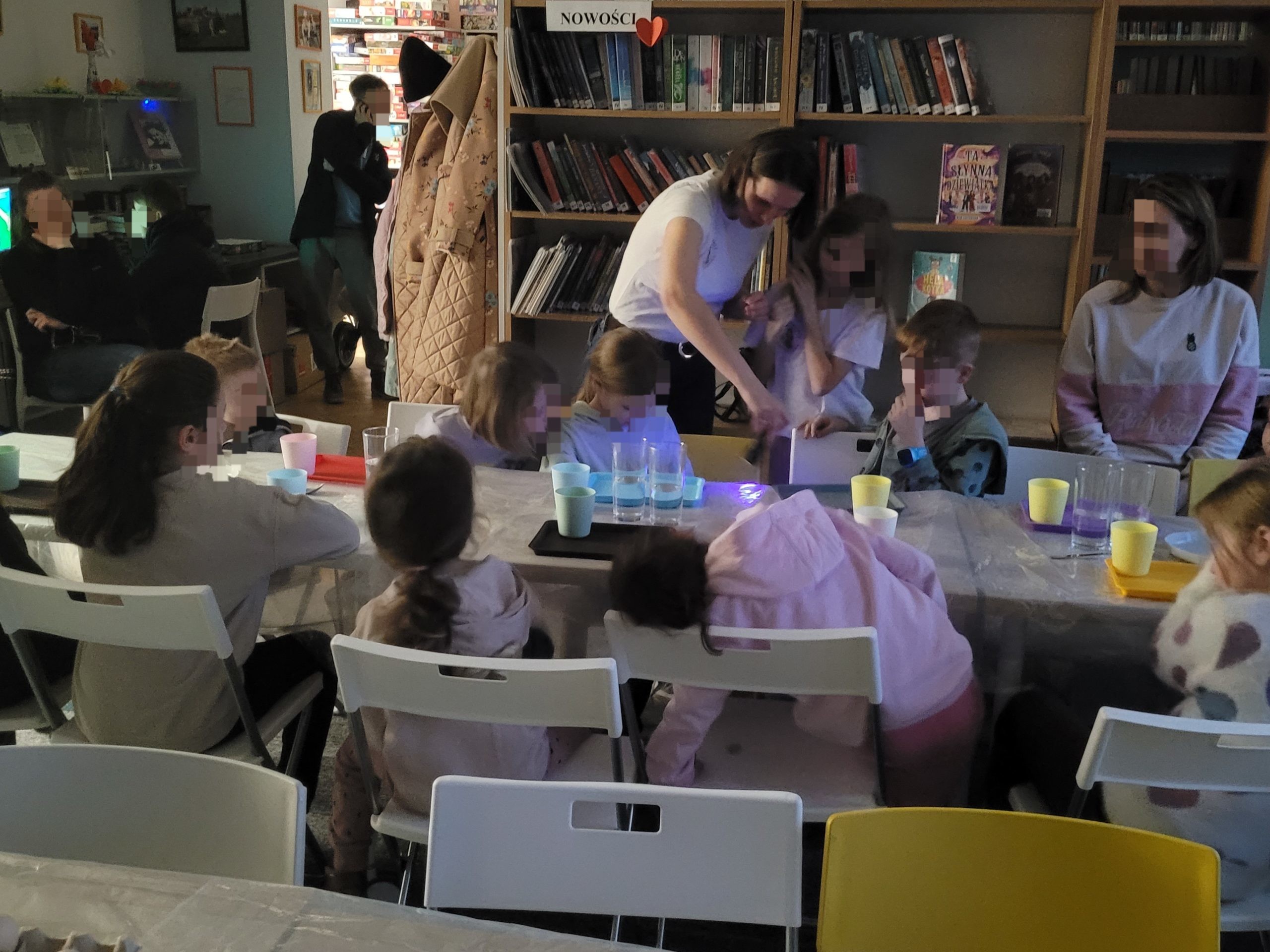 Dzieci siedzą naprzeciw siebie przy stole i słuchają prowadzącej, która tłumaczy przebieg eksperymentu. Na stole stoją kubki i szklanki z wodą.