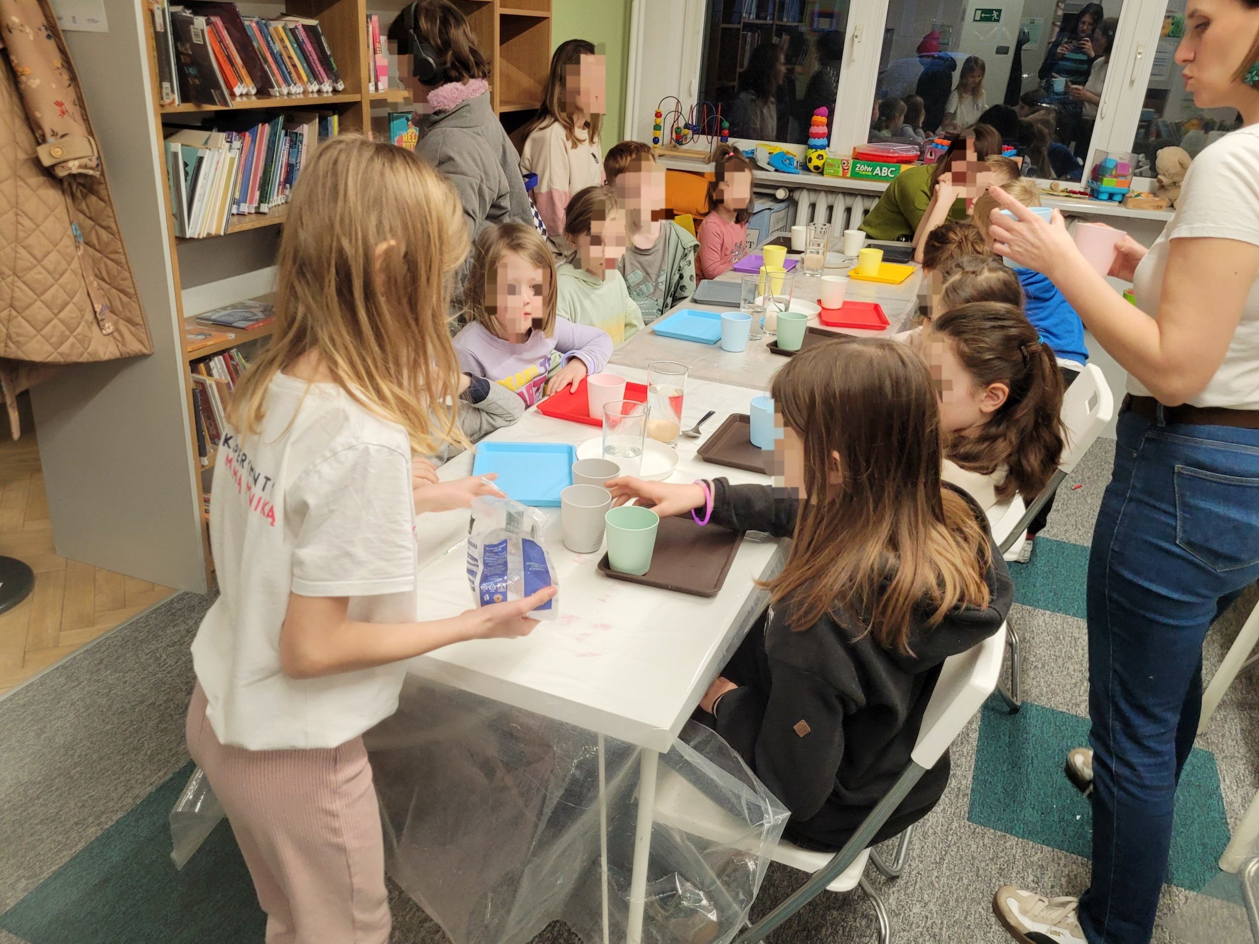 Dzieci siedzą przy długim stole w bibliotece i biorą udział w warsztatach edukacyjnych z eksperymentami, używając kubków, tacek i jajek. Zajęcia prowadzi dorosła osoba.