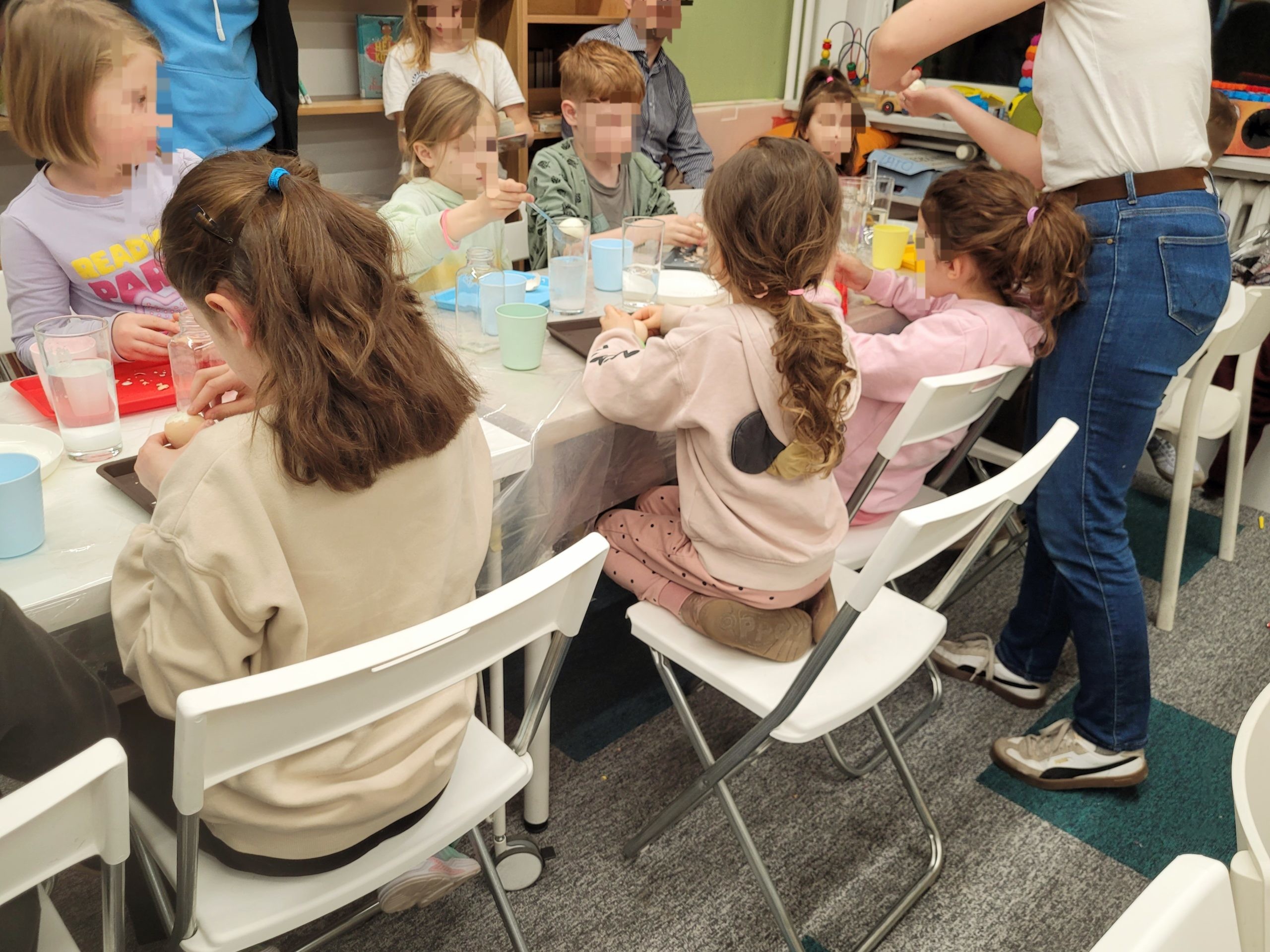 Widok z boku na dzieci siedzące przy stole podczas warsztatów w bibliotece. Na stole znajdują się naczynia, kubki i materiały do eksperymentów.
