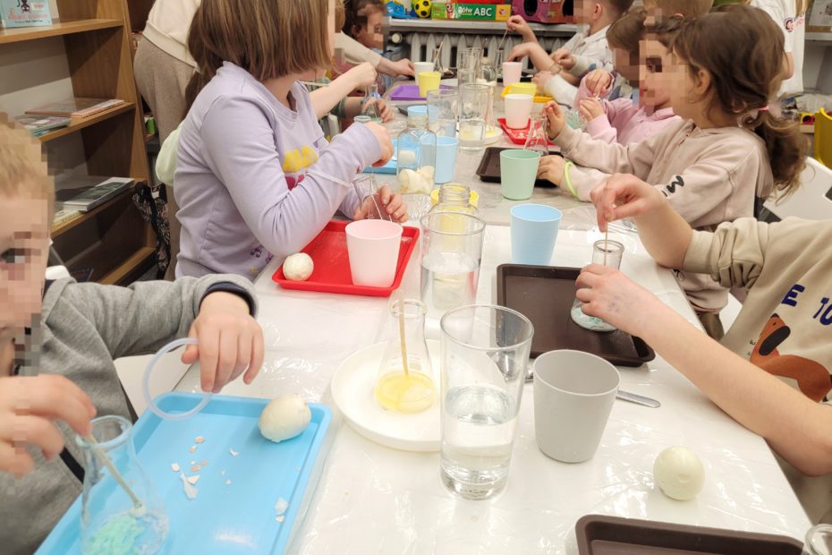 Uczestnicy warsztatów – dzieci w różnym wieku – wykonują doświadczenia przy stole, mieszając substancje w naczyniach i obserwując efekty.