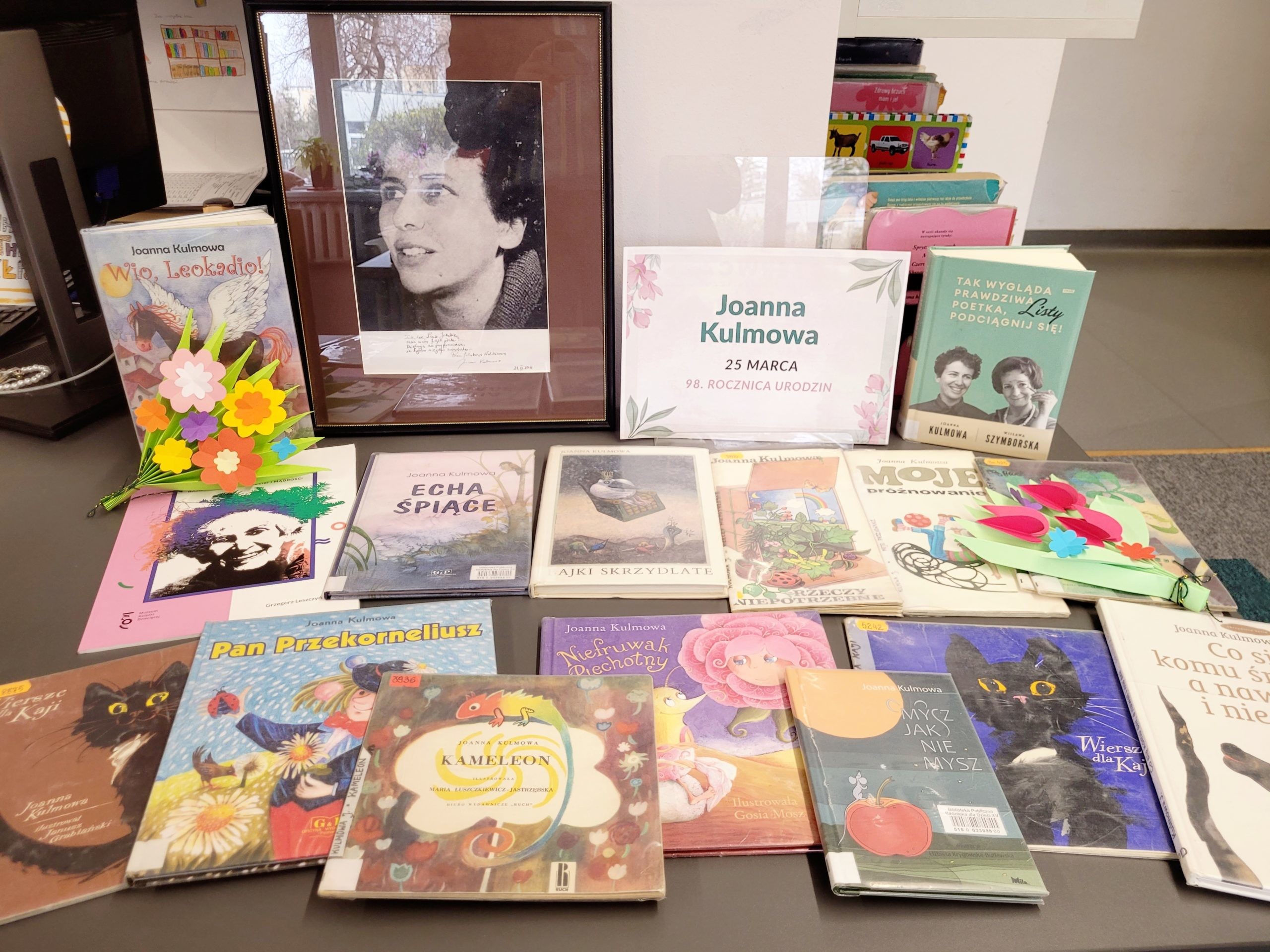 Ekspozycja w bibliotece poświęcona Joannie Kulmowej: portret poetki, informacja o 98. rocznicy urodzin oraz książki jej autorstwa ułożone na stole.