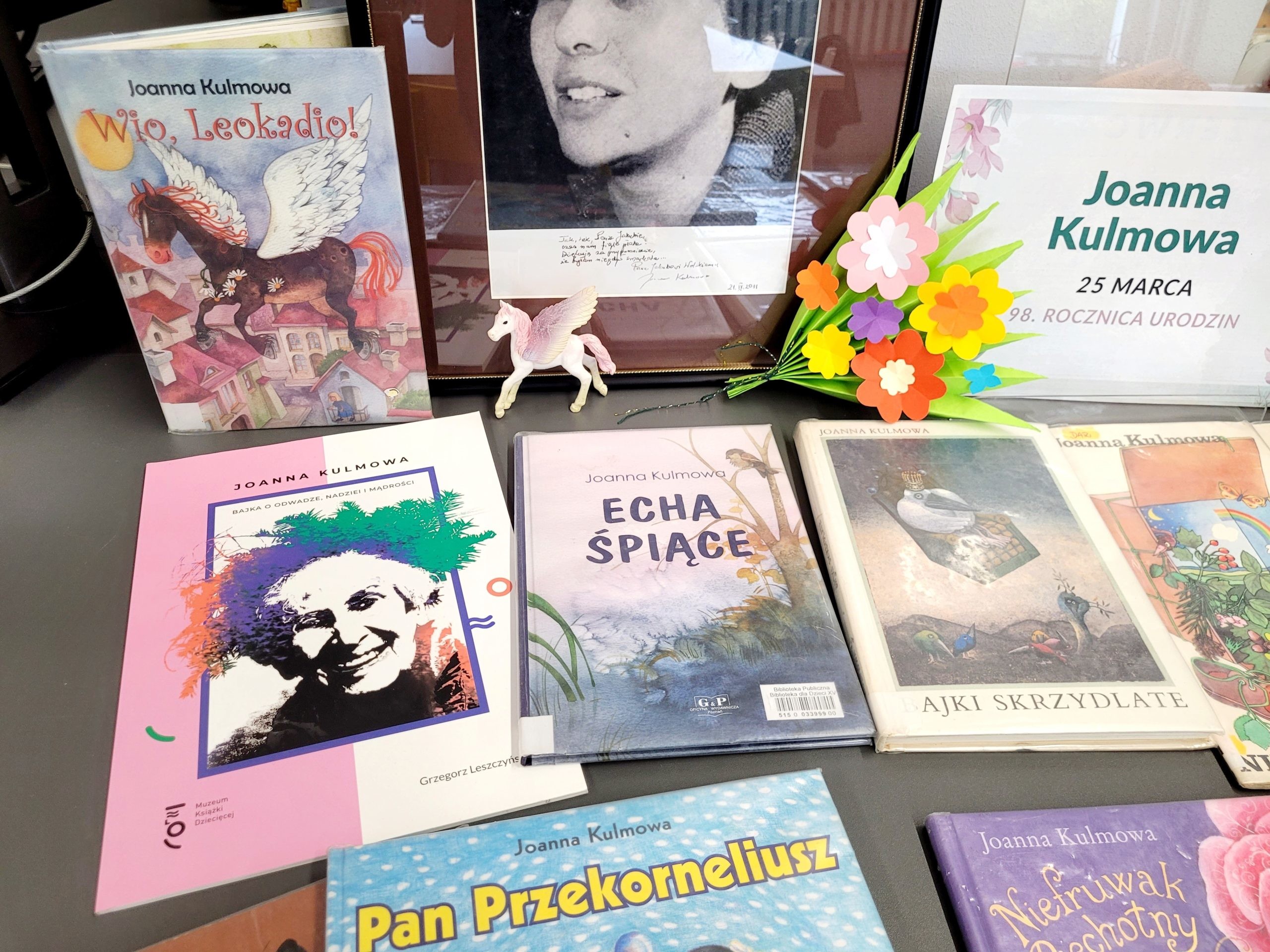Zbliżenie na wystawę książek Joanny Kulmowej oraz kartę informacyjną z datą urodzin, ozdobioną papierowymi kwiatami wykonanymi przez dzieci.