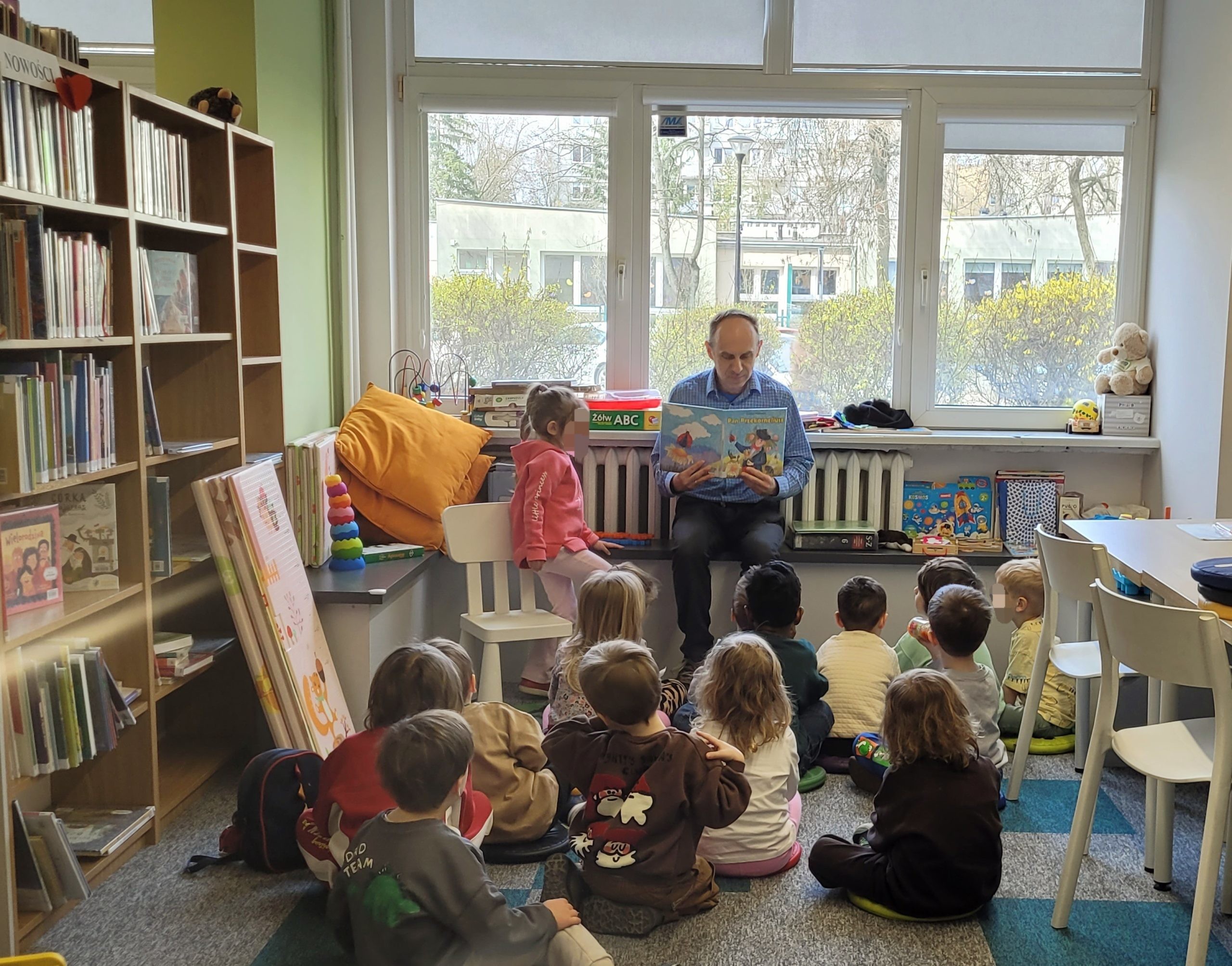 Bibliotekarz czyta książkę dzieciom siedzącym na dywanie w sali bibliotecznej, w tle regały z książkami i okno.