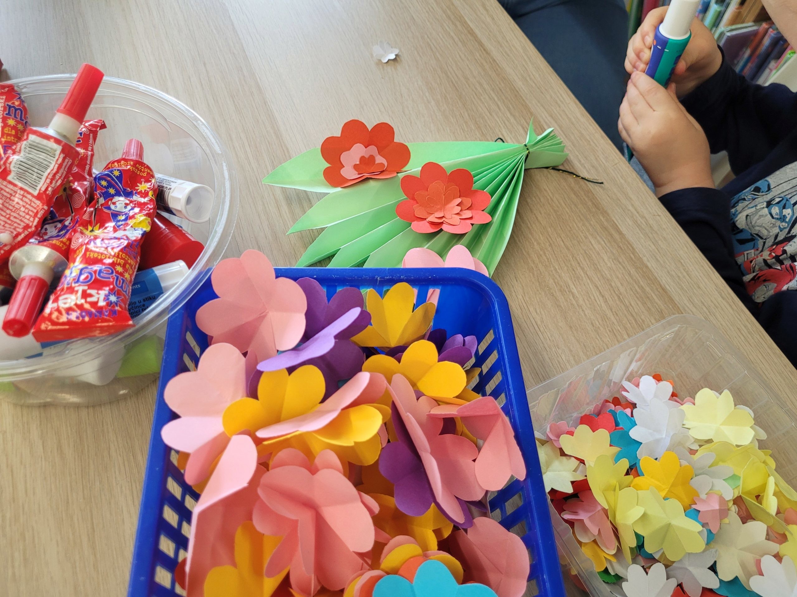 Materiały plastyczne na stole: papierowe kwiaty, kleje oraz elementy przygotowane do pracy plastycznej.