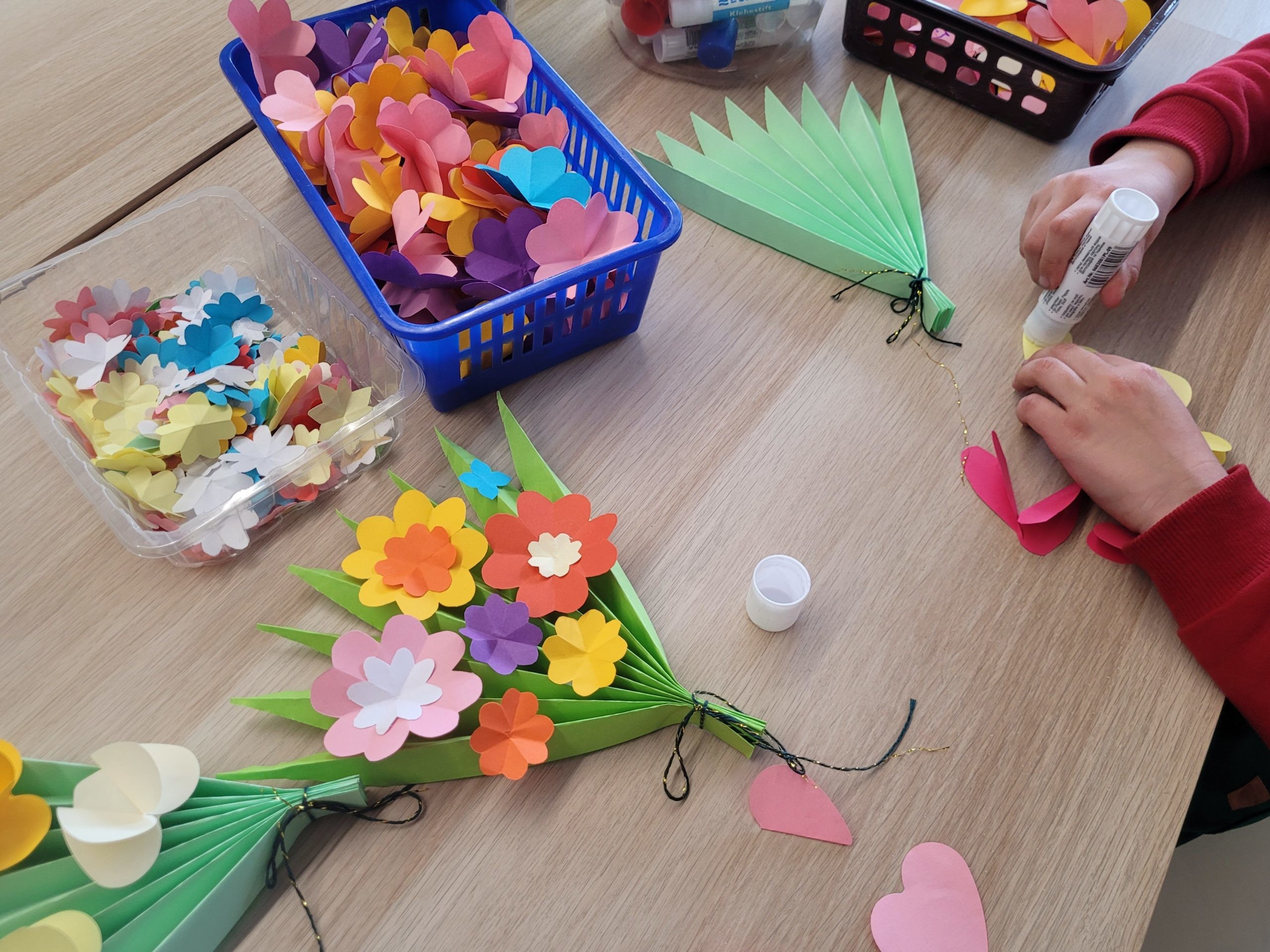 Dziecięce ręce przyklejają papierowe kwiaty do zielonej, składanej formy przypominającej bukiet.