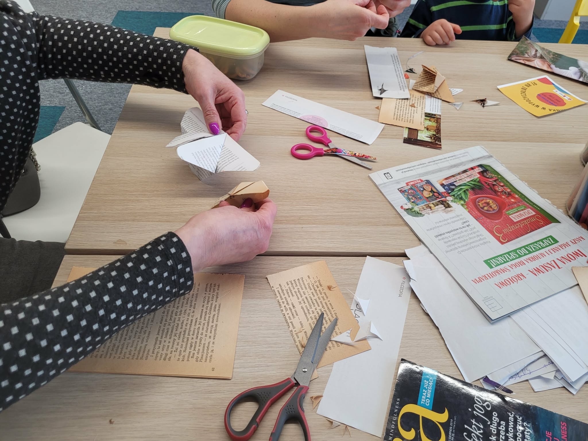 Dłonie dorosłej osoby i dziecka podczas składania papierowego motyla przy stole; na blacie widoczne nożyczki, wycinki z książek i papier.