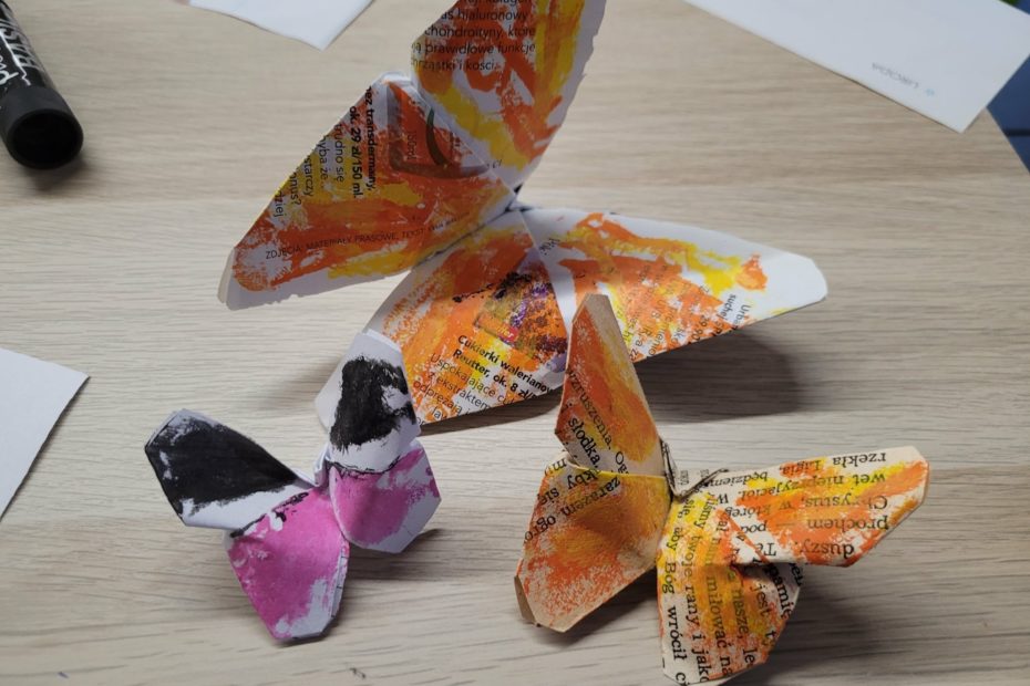 Kilka trójwymiarowych papierowych motyli stojących na stole; wykonane z papieru z nadrukiem i pomalowane farbami w odcieniach pomarańczowym, różowym i czarnym.