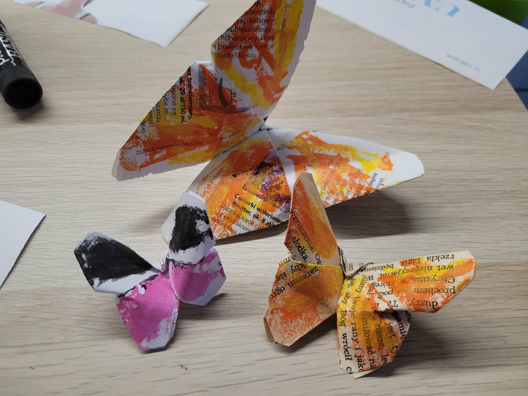 Kilka trójwymiarowych papierowych motyli stojących na stole; wykonane z papieru z nadrukiem i pomalowane farbami w odcieniach pomarańczowym, różowym i czarnym.