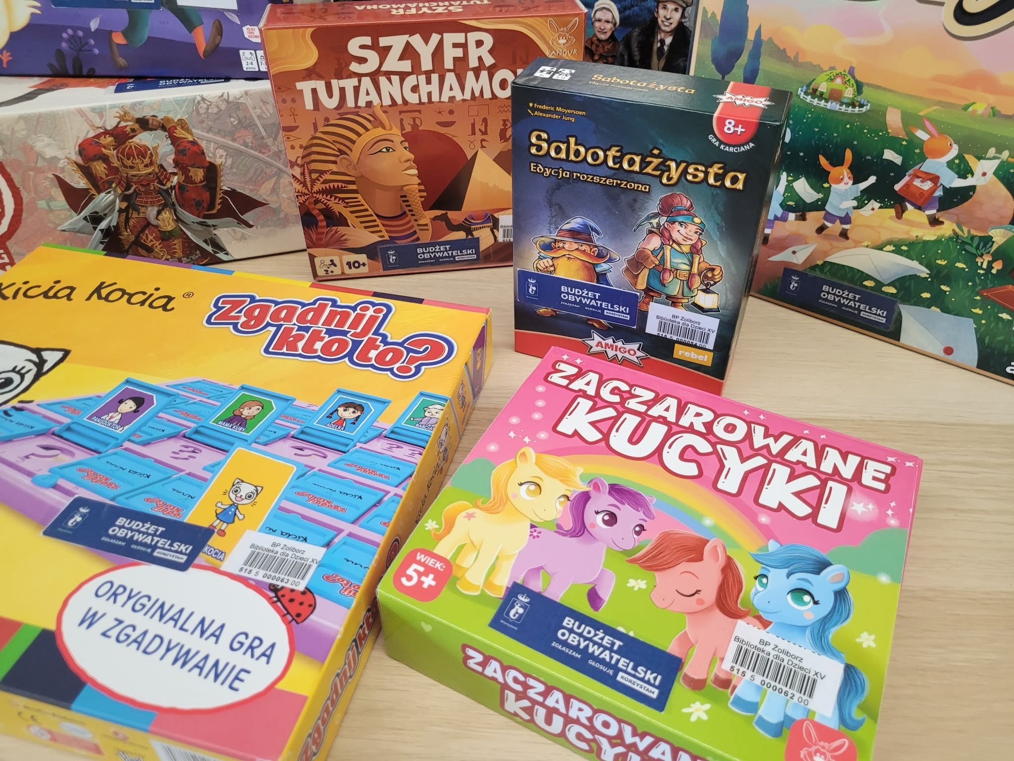 Zestaw kolorowych gier planszowych dla dzieci i rodzin ułożonych na stole, m.in. „Zgadnij kto to?”, „Zaczarowane kucyki”, „Sabotażysta” oraz „Szyfr Tutanchamona”.