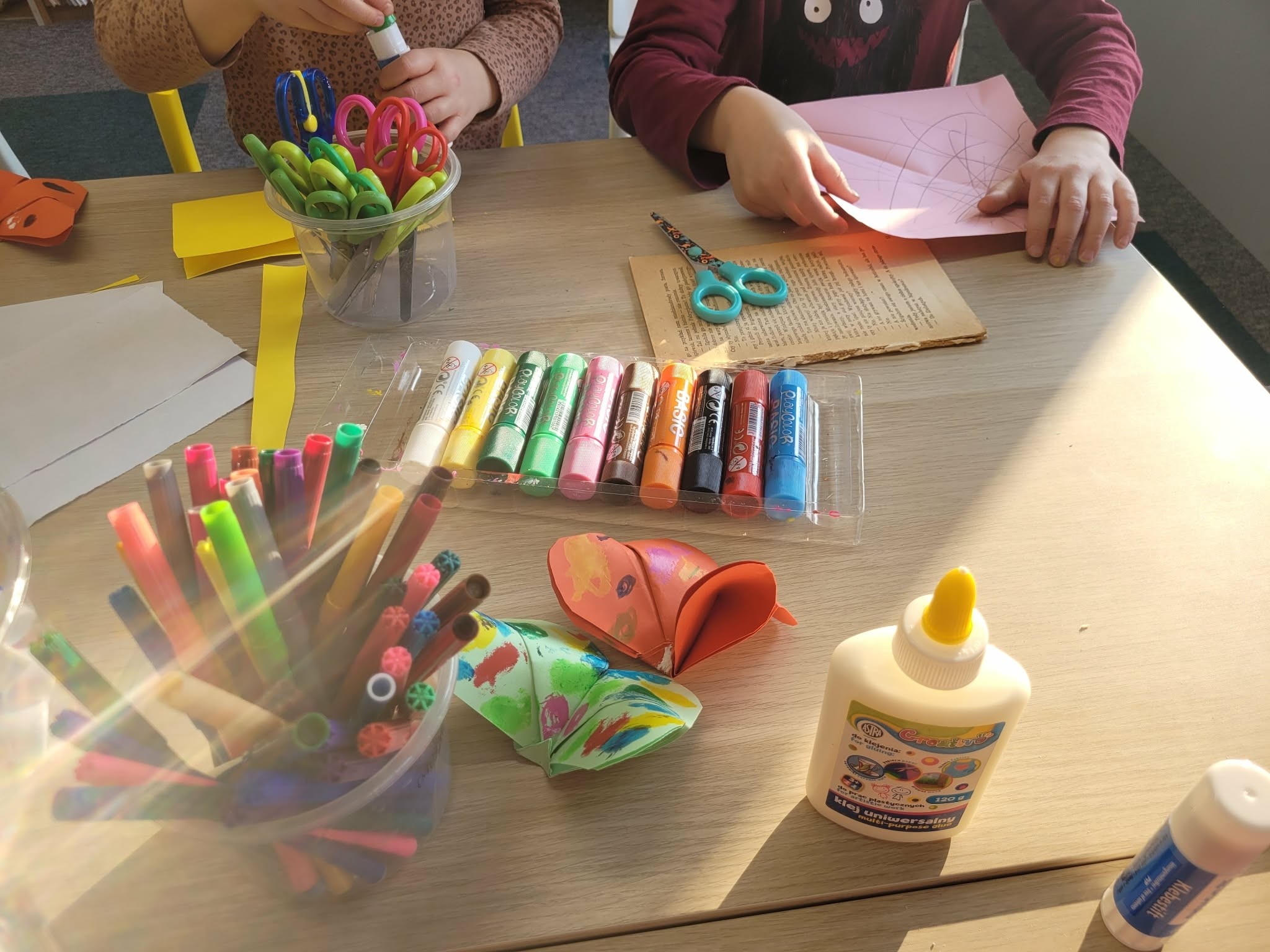 Dzieci przy stole warsztatowym wykonują prace plastyczne; na blacie kolorowe flamastry, klej, nożyczki oraz gotowe papierowe motyle.