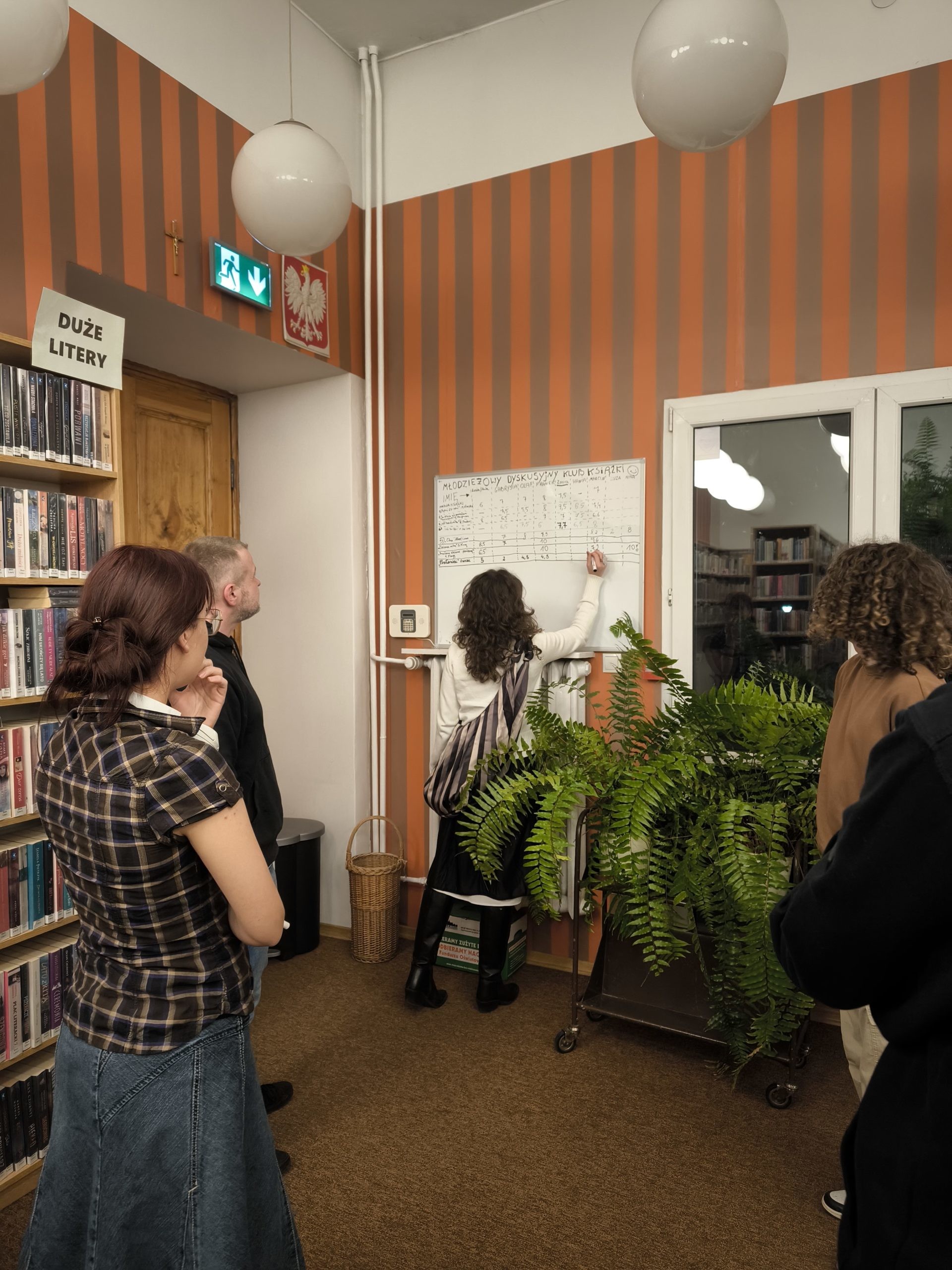 grafika przedstawiająca członków dyskusyjnego klubu książki podczas zapisywania ocen na tablicy po omówieniu książki "Posłaniec" Markusa Zusaka